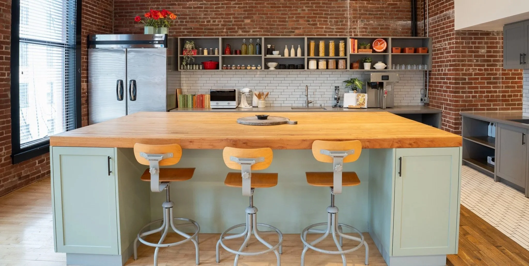 Modern kitchen with brick walls, a large wooden island with cream cabinets, four wooden barstools, open shelving with jars and dishes, a stainless steel refrigerator, and a window with blinds.