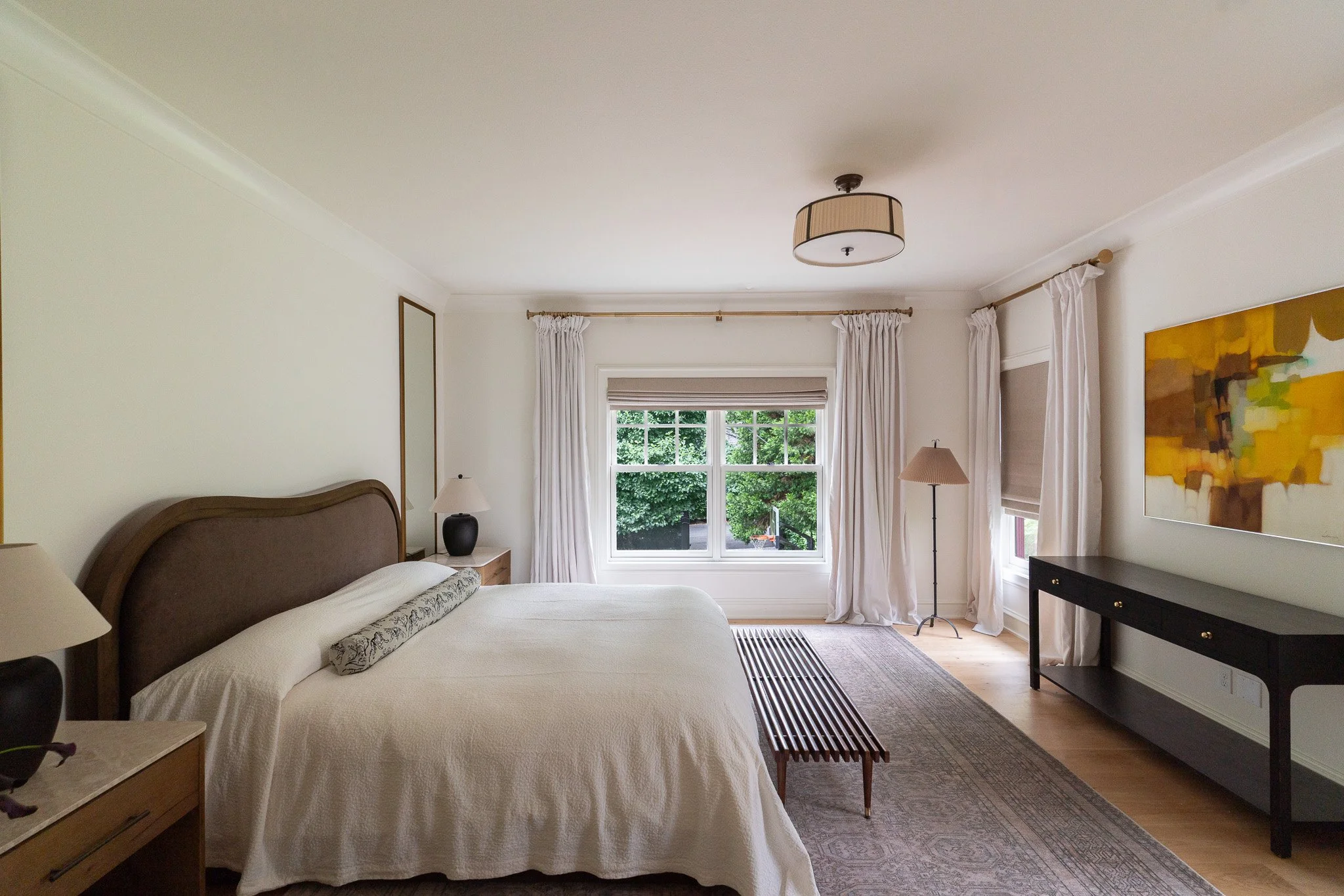 A bedroom with a large bed, nightstand with a lamp, window with white curtains, and abstract artwork on the wall.