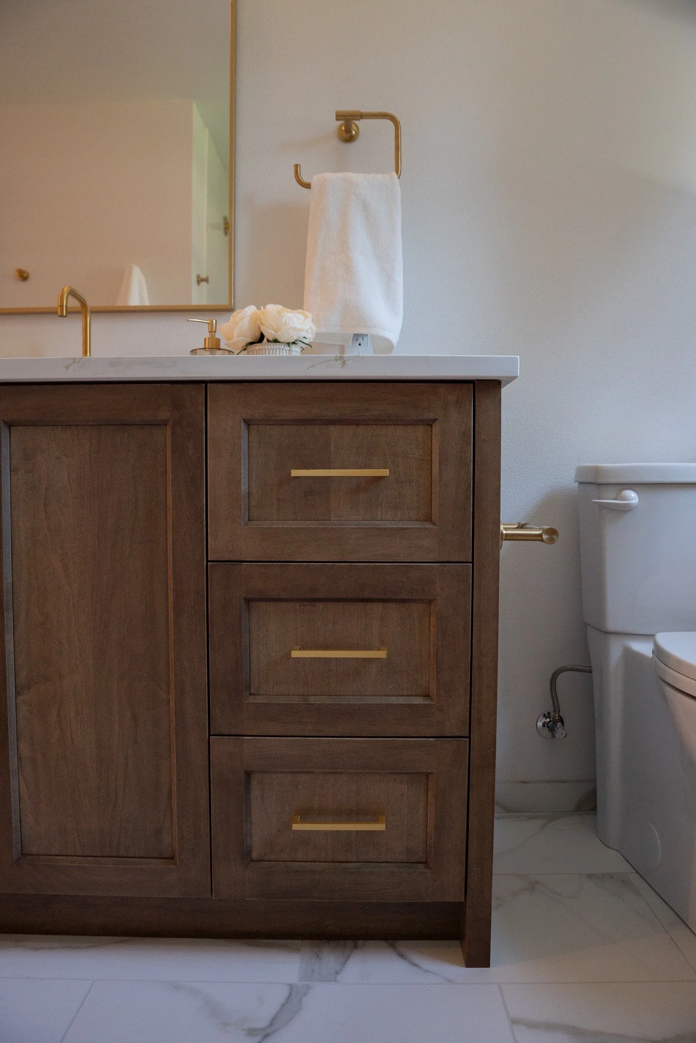 Bathroom vanity with a wood cabinet, white marble countertop, and gold hardware. A mirror, white towel on a gold towel holder, and a small vase with white flowers sit on the counter. Part of a toilet is visible on the right. The floor is white marble