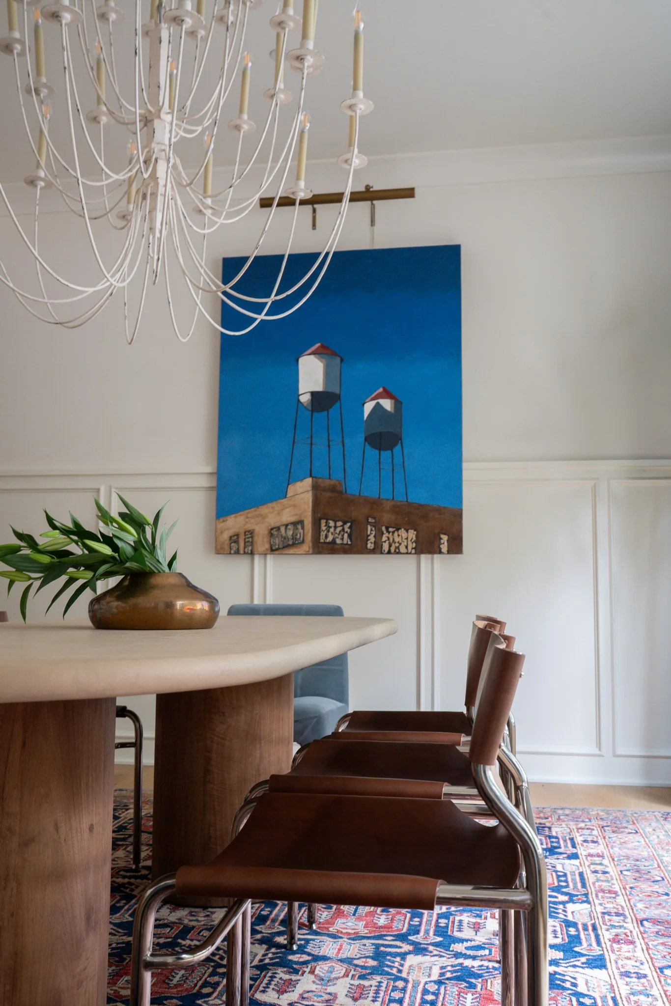 A dining room with a round wooden table, potted plant, colorful patterned rug, chairs with brown leather seats, artwork of water towers on the wall, and a white chandelier hanging from the ceiling.