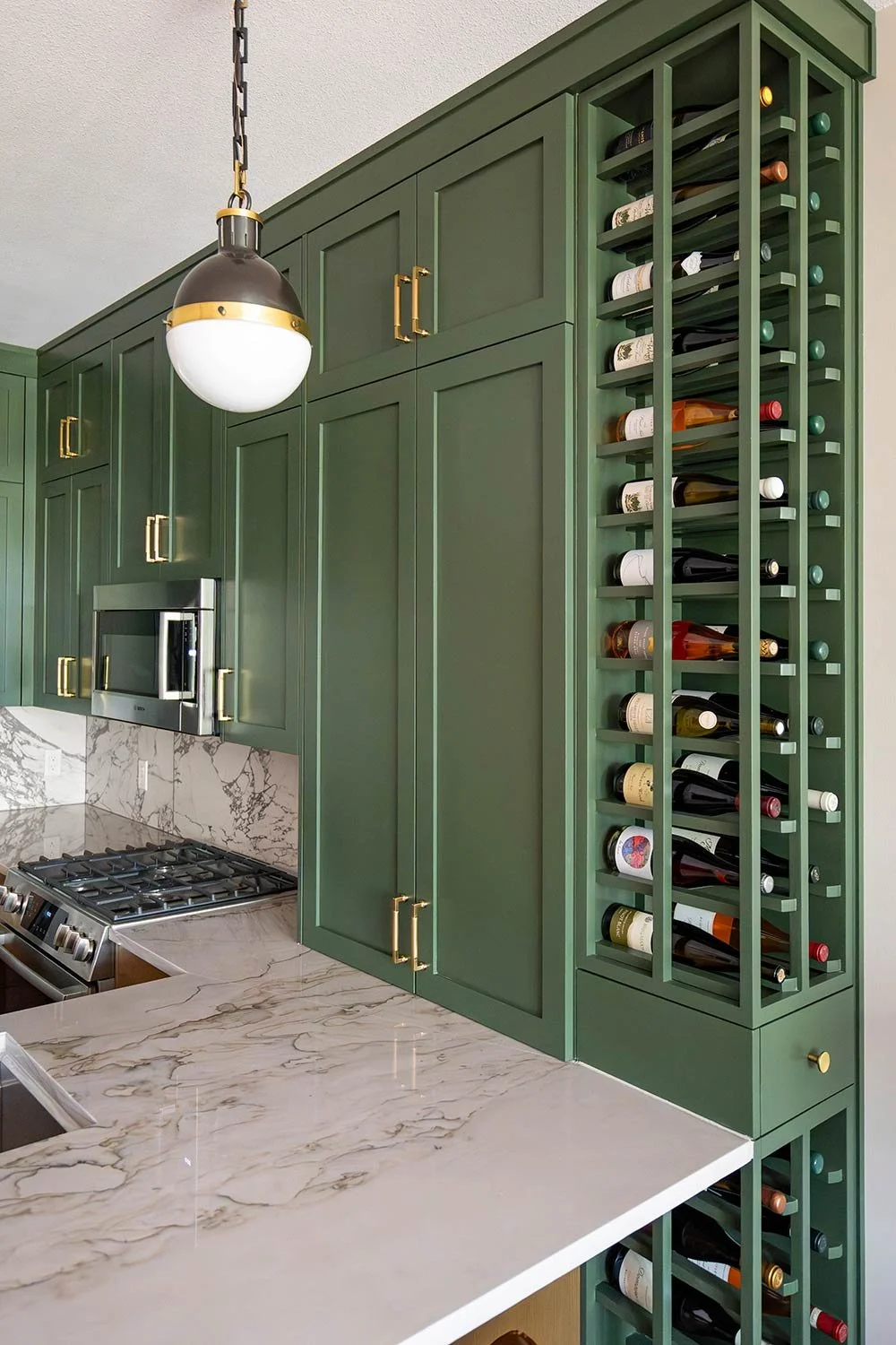 A kitchen with green cabinets, marble countertops, and a built-in wine rack filled with wine bottles, along with a black and white hanging light fixture.