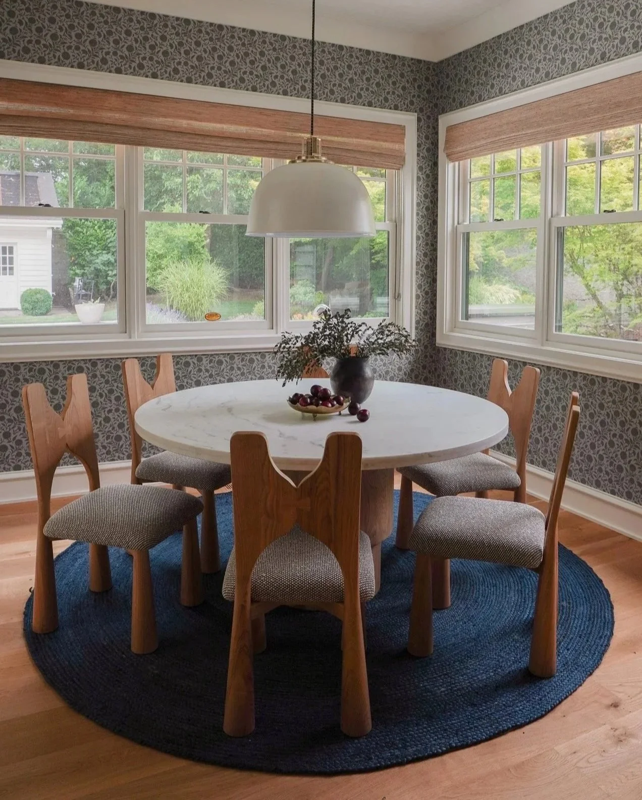 A cozy dining area with a round white marble table, surrounded by six wooden chairs with upholstered seats. On the table, there is a black vase with greenery and a bowl of red grapes. The area has large windows with light brown shades, showing a view of a lush green backyard. The floor is wooden, and a dark blue round rug is underneath the table.