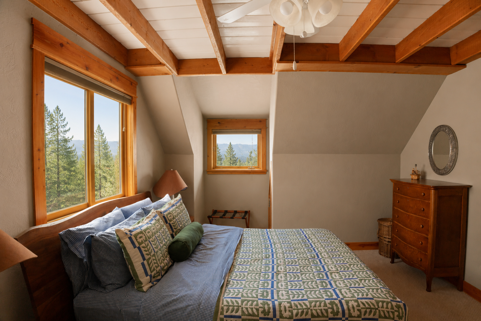 A cozy bedroom with a wooden bed frame, patterned bedding and pillows, and a window showing a view of trees and mountains. There's a small window, a dresser with a round mirror, and a basket in the corner. The ceiling has wooden beams and a ceiling fan.