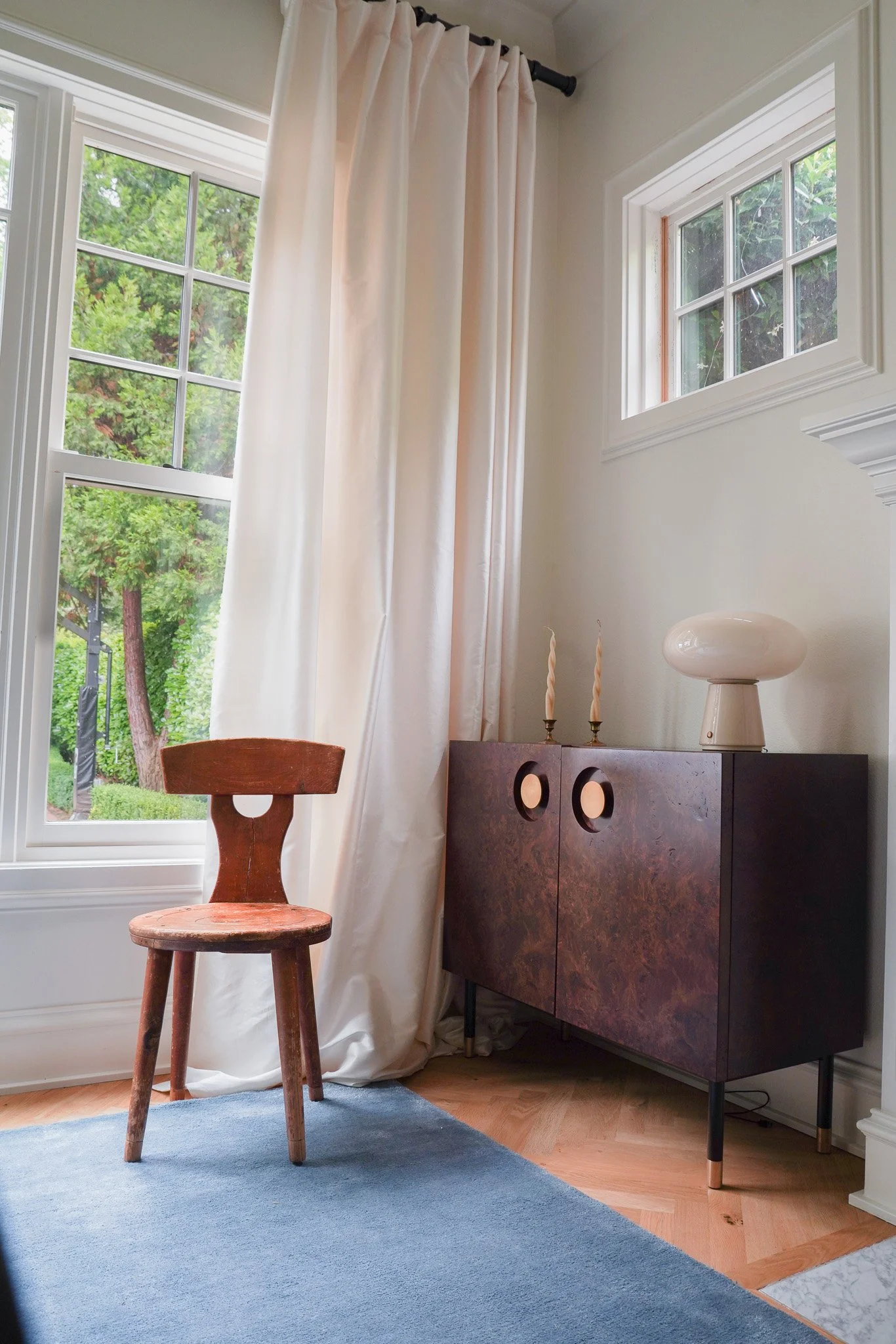 A room corner with a wooden chair in front of a window with white curtains and a cabinet with decorative items on top, including two twisted candles and a white lamp. The room has natural light coming in through the windows, and there is a blue rug o