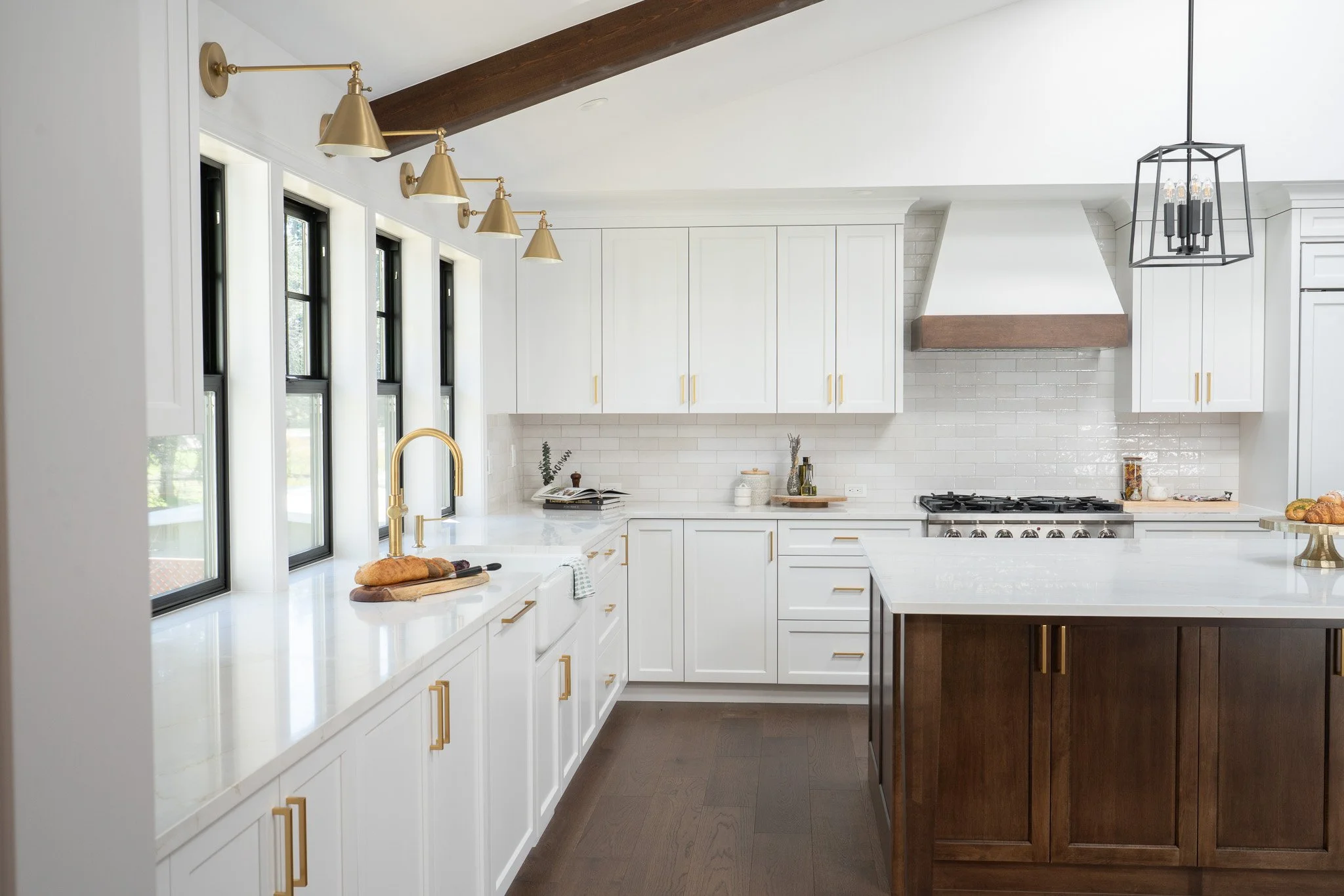 A modern kitchen with white cabinets, black-framed windows, a white countertop, and a dark wooden island. Gold fixtures include a faucet and cabinet handles. Two door-style pendant lights and a black open-frame chandelier hang from the ceiling. Items