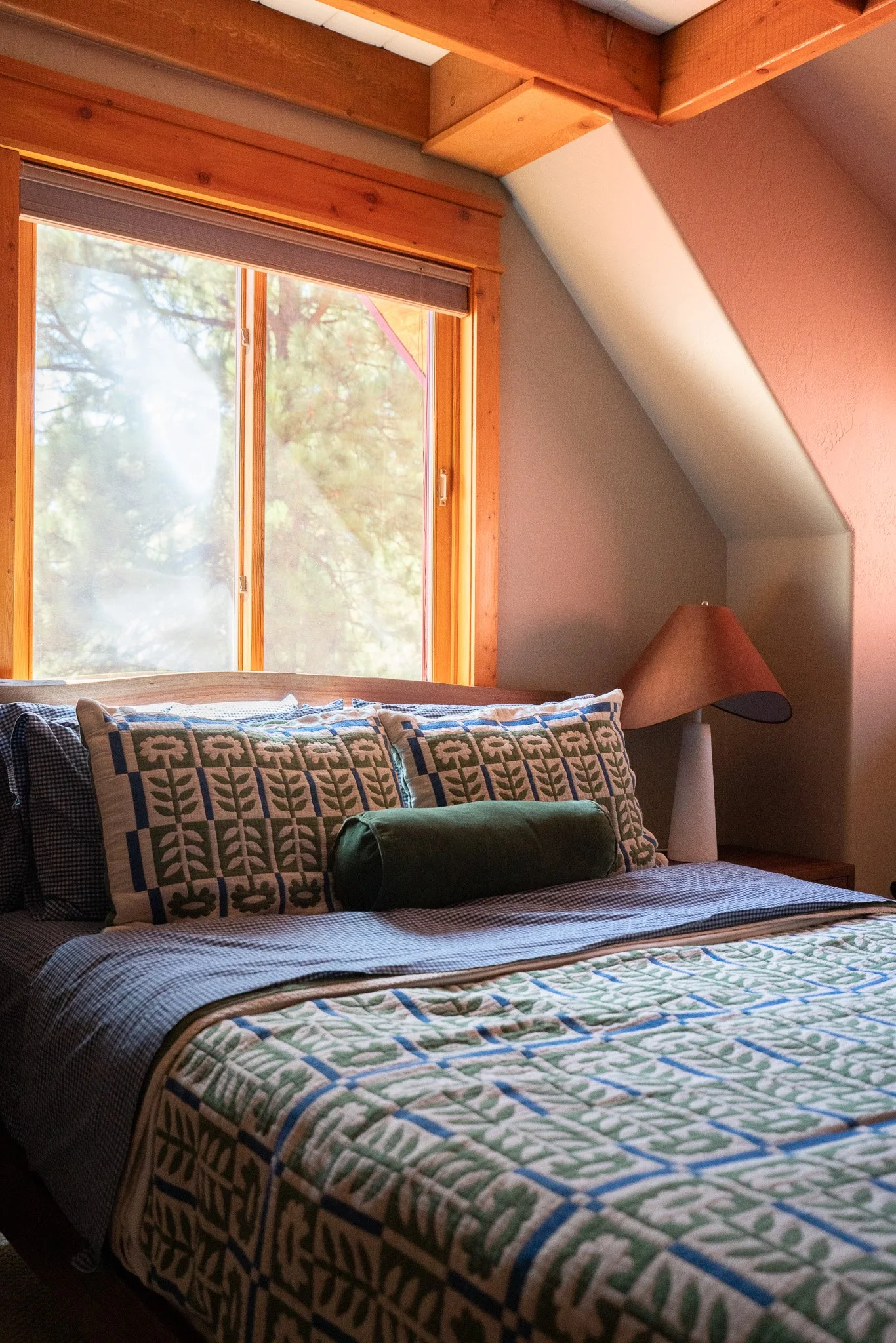 A cozy bedroom with a large window letting in natural light, showcasing a bed with a patterned quilt and pillows, and a side table with a lamp.
