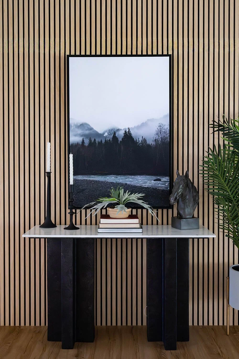 A decorative table with two candles, a potted succulent plant, a stack of books, and a horse head sculpture in front of a window showing a mountainous landscape with fog and trees, set against a wooden slat wall.