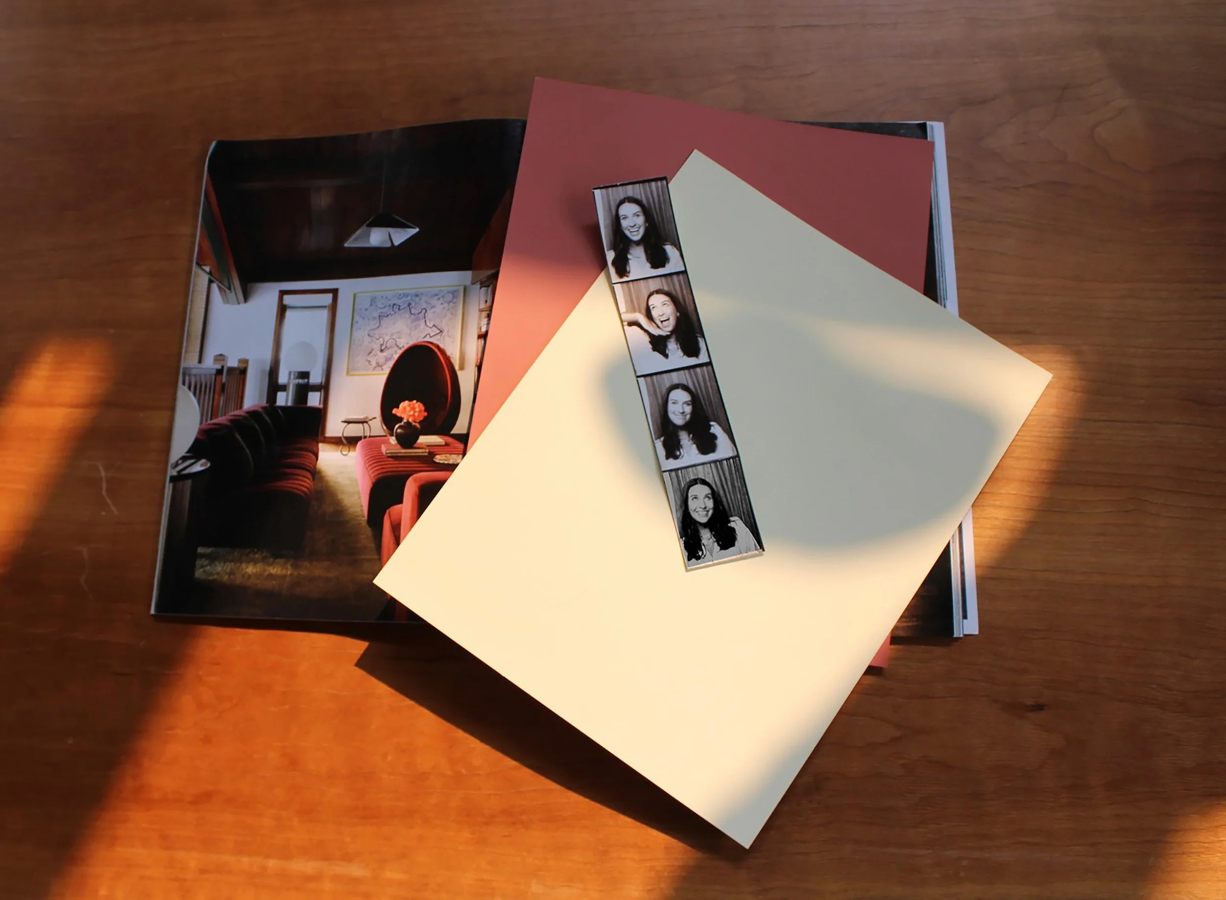 Open magazine with interior decor photo, photo booth strip with smiling woman, and blank colored papers on a wooden table.