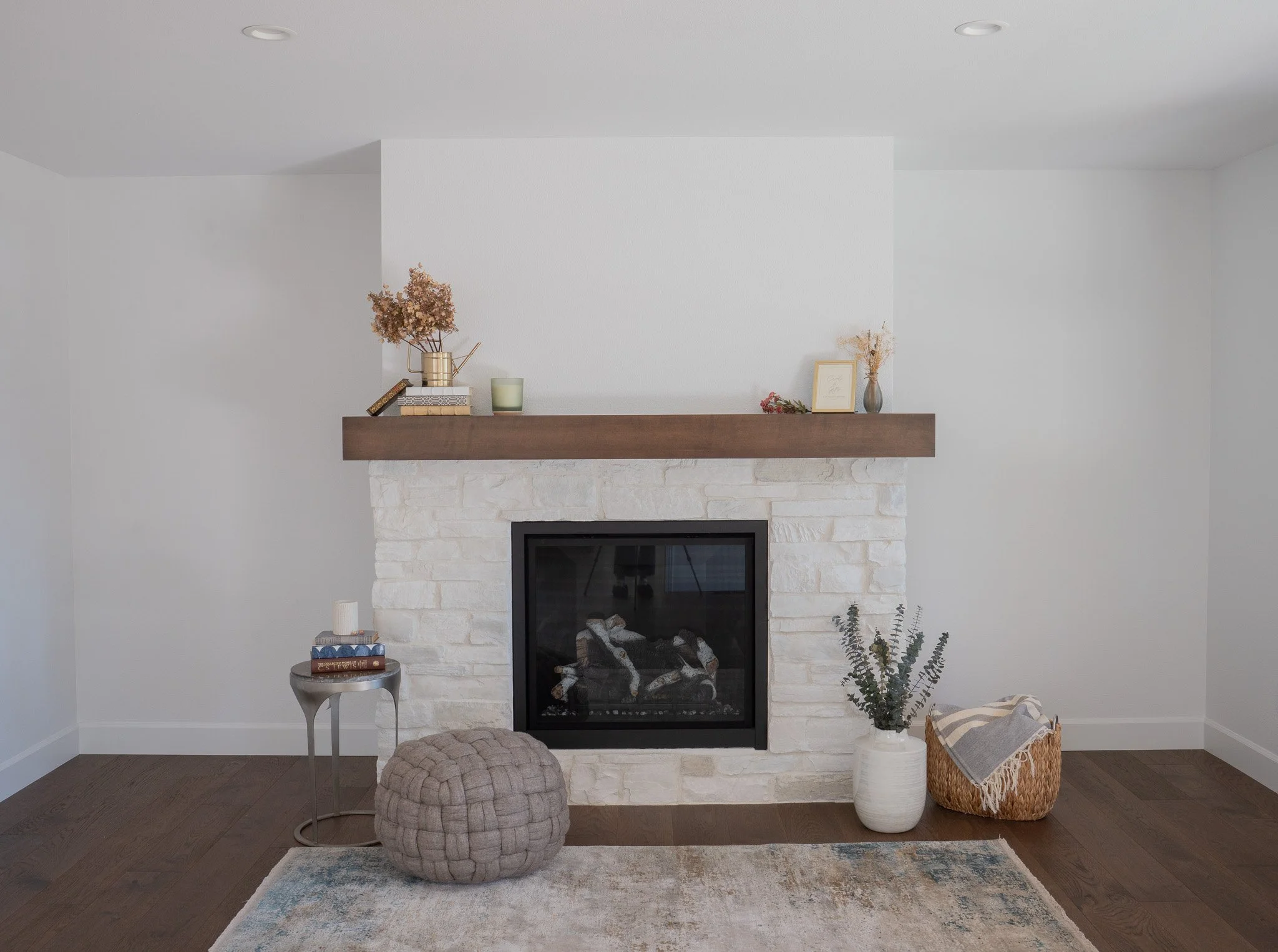 Living room with a stone fireplace and wooden mantel decorated with dried flowers, a framed photo, and books. A side table with books, a small plant, and a candle. In front, a beige knotted pouf. To the right, a white vase with dried plants and a bas