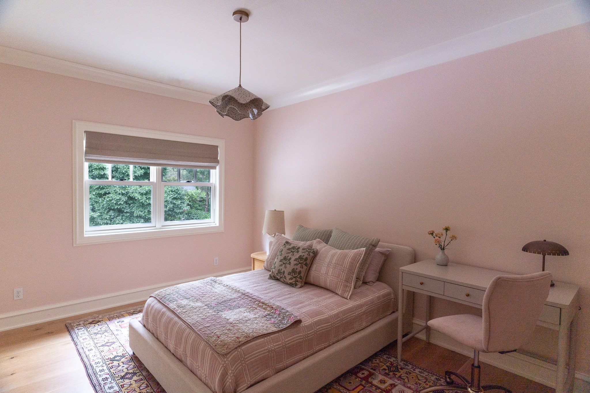A bedroom with pink walls, a window with a gray blind, and a bed with a floral quilt and pillows. There is a white nightstand with a vase of flowers and a lamp, and a beige chair. The ceiling has a pendant light fixture.