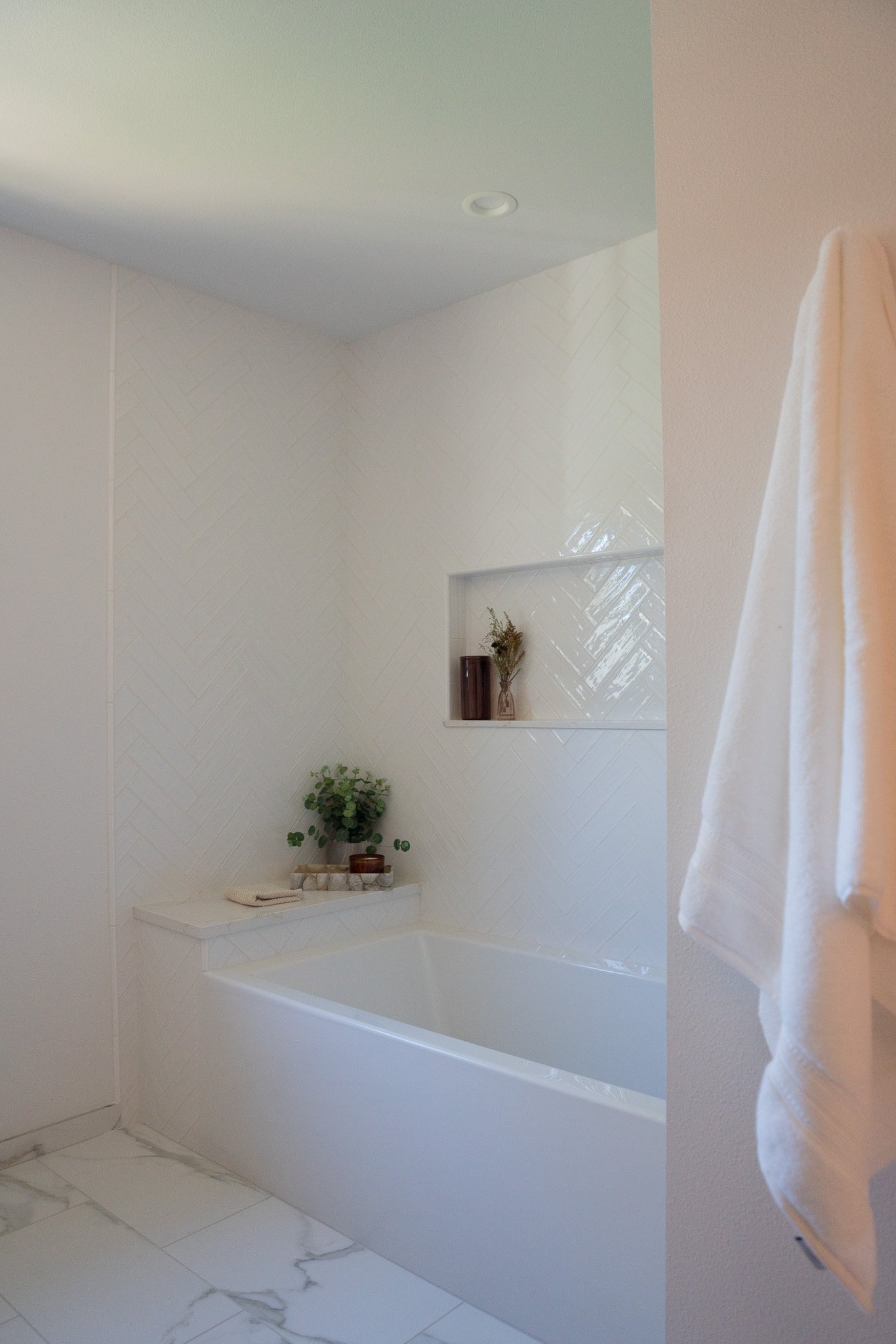 A bathroom with a white bathtub, a small white shelf holding a potted plant, a folded towel, and decorative items, beige wall tiles, an inset shelf with vases, and a towel hanging on a hook.