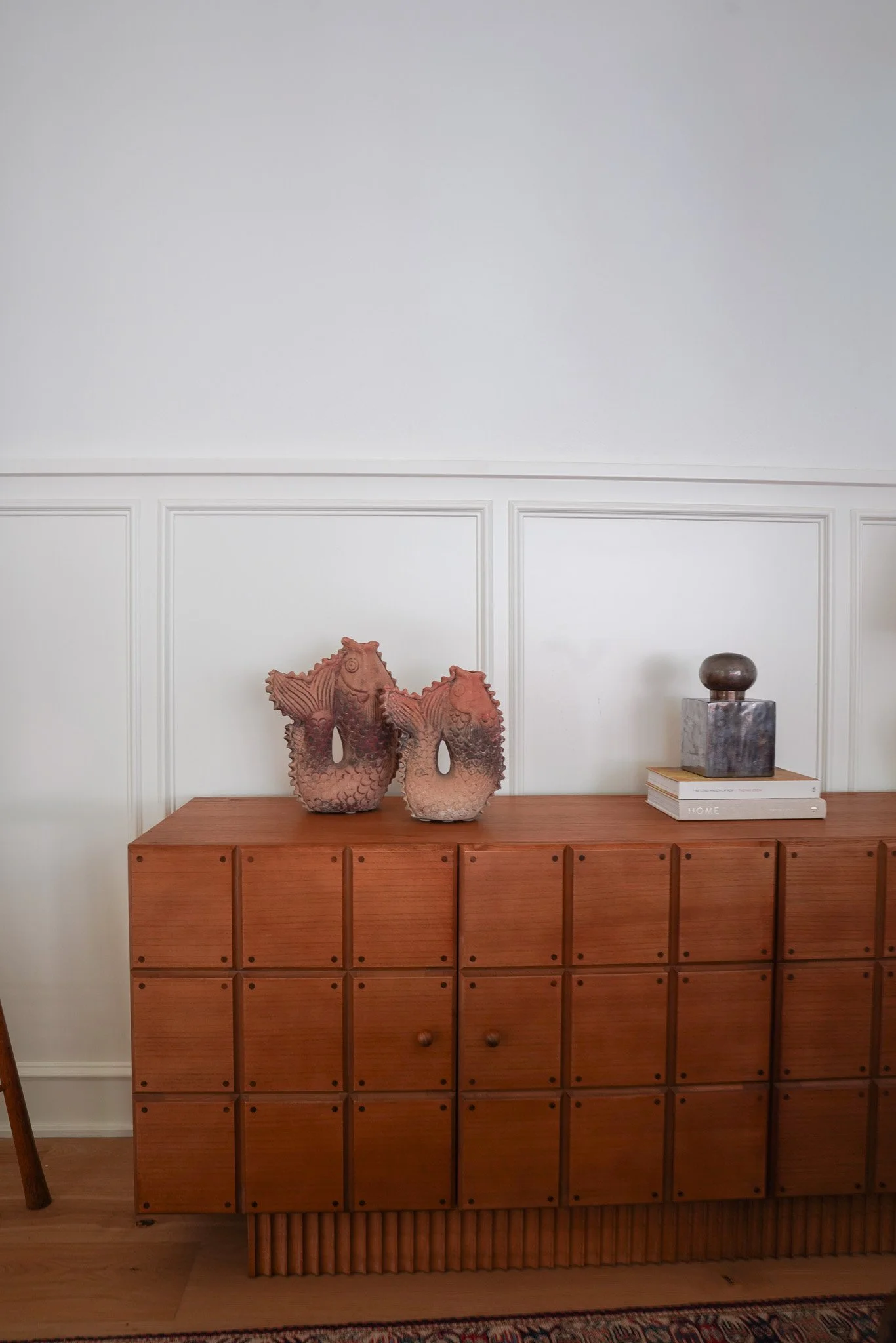 A mid-century wooden sideboard with multiple small drawers, placed against a white wall with decorative wall molding. On top, there are two sculptural fish-shaped ceramic vessels, a marble sculpture of a sphere on a square base, and a stack of two bo