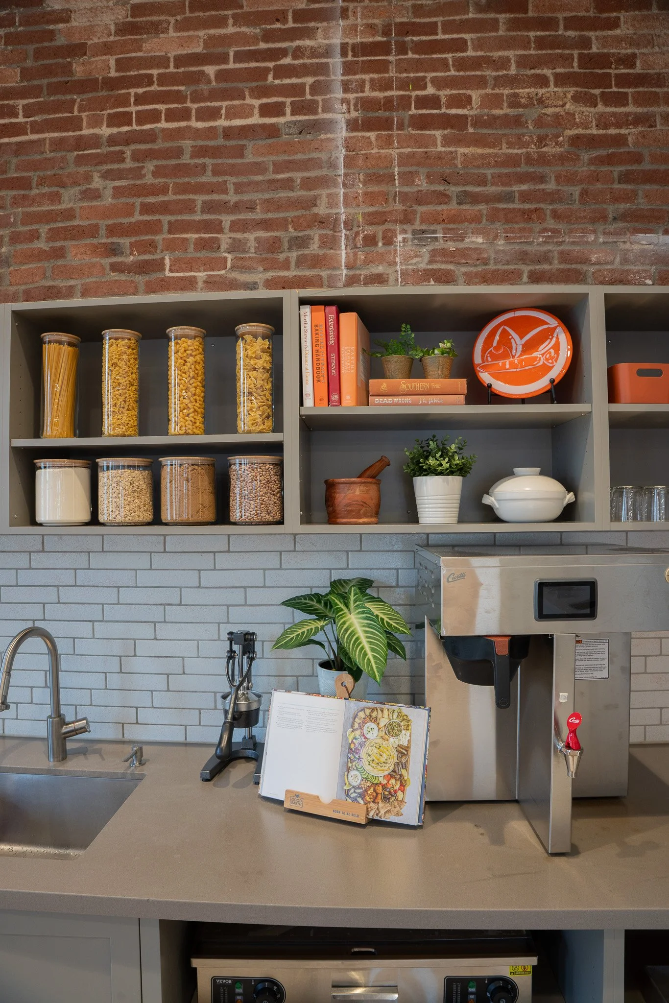 A kitchen counter with a sink, a potted plant, a menu, and coffee machine. Above, a gray shelf with jars of pasta, books, a plate with utensils, and a small plant. The background features a exposed brick wall.