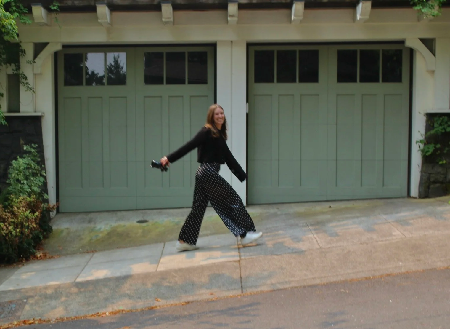 Woman with long hair smiling, walking on sidewalk in front of a light green garage door, holding an item in her right hand, wearing a black jacket, black and white patterned pants, and white shoes.