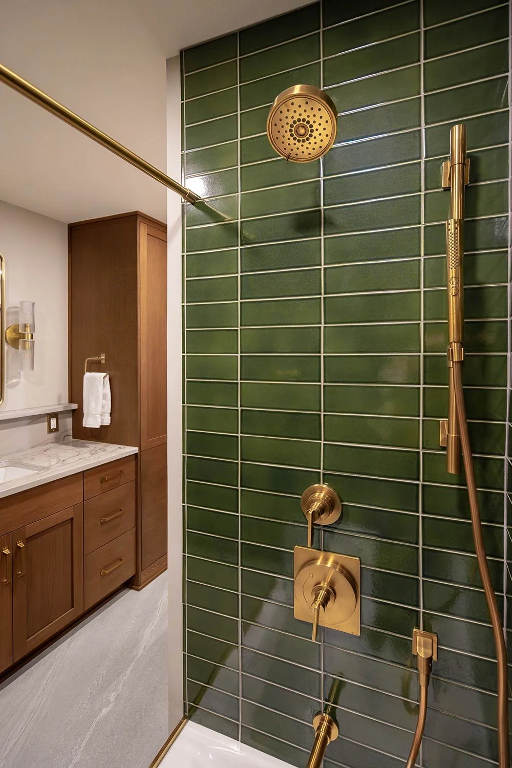 Close-up of a modern bathroom shower with dark green tiles, gold fixtures, and a handheld showerhead. Part of a wooden vanity with a white marble countertop and a towel rack is visible in the background.