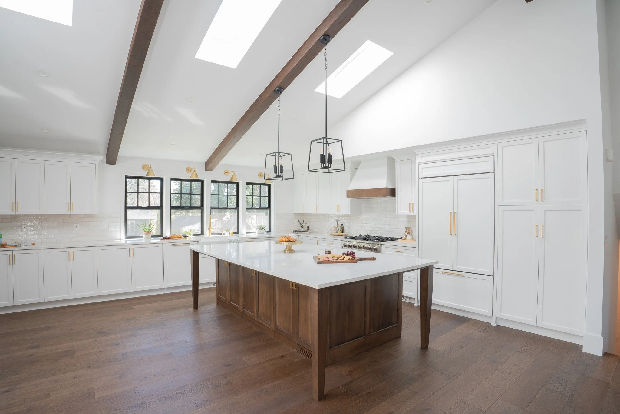Bright and spacious kitchen with white cabinets, black-framed windows, and a large wooden kitchen island with a white countertop. Skylights and hanging pendant lights provide natural and artificial lighting. Decor includes small plants and food on th