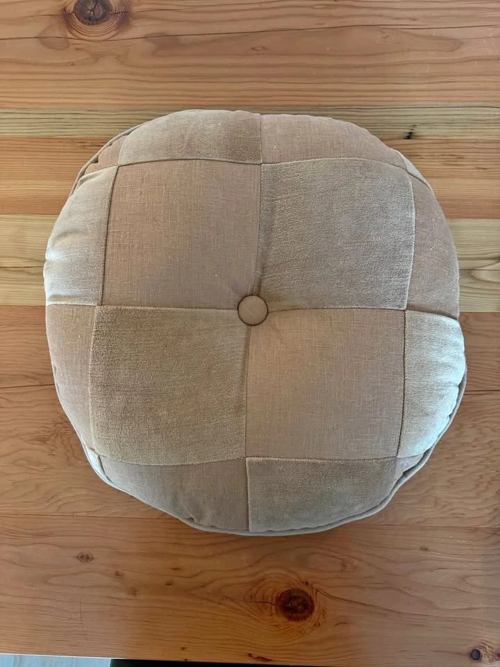A round, beige, fabric ottoman with a tufted top and a button in the center, placed on a wooden floor.