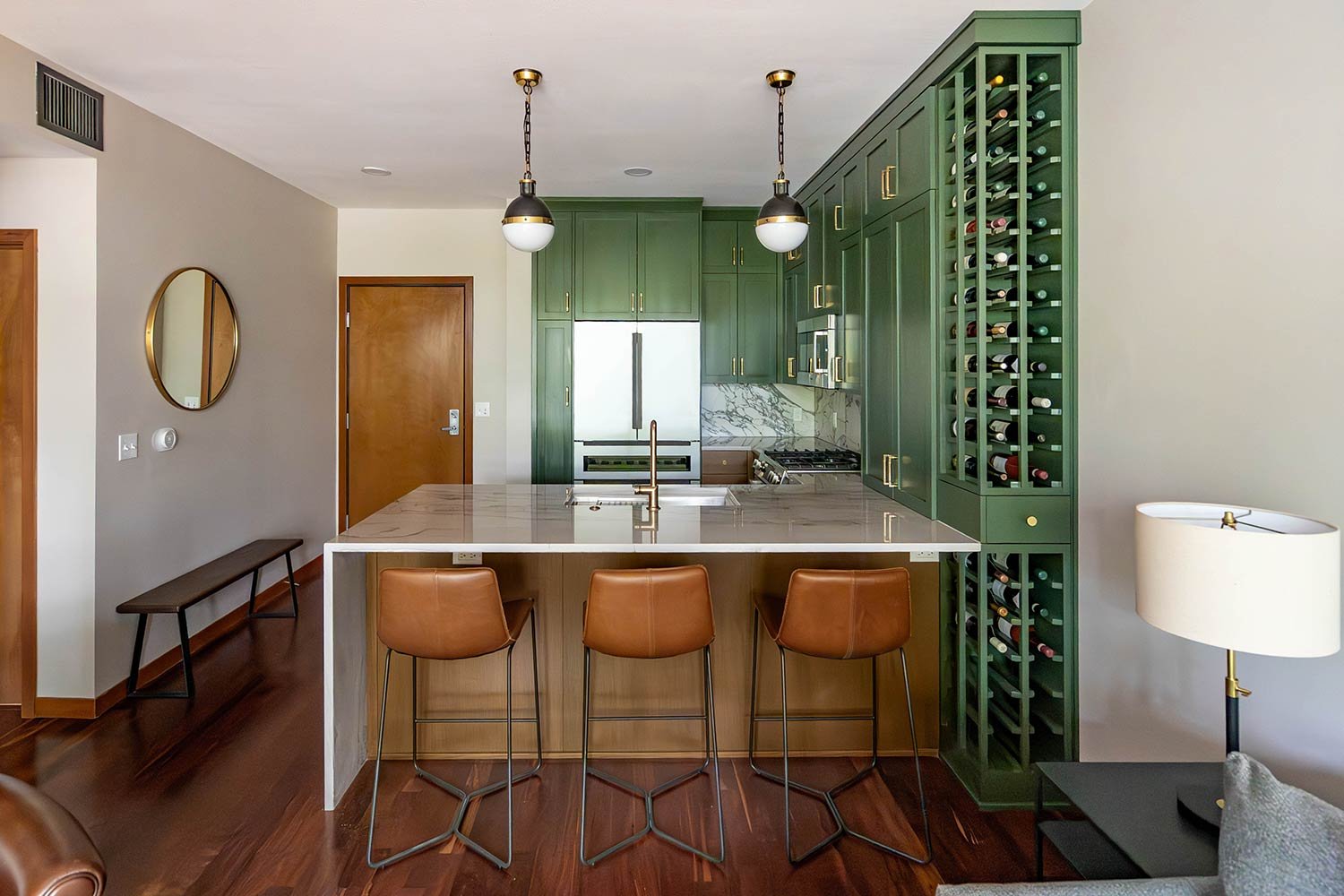 Kitchen with green cabinets, marble countertop island, three tan barstools, wine storage, stainless steel refrigerator, and a modern chandelier.