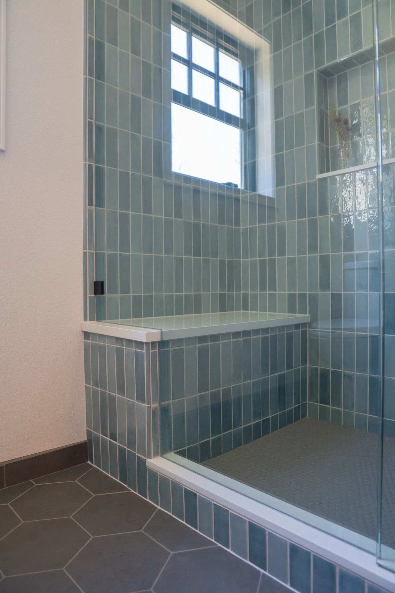 Shower area with blue tiles on walls and grey tiles on floor, enclosed by a glass door and wall, with a window letting natural light in.