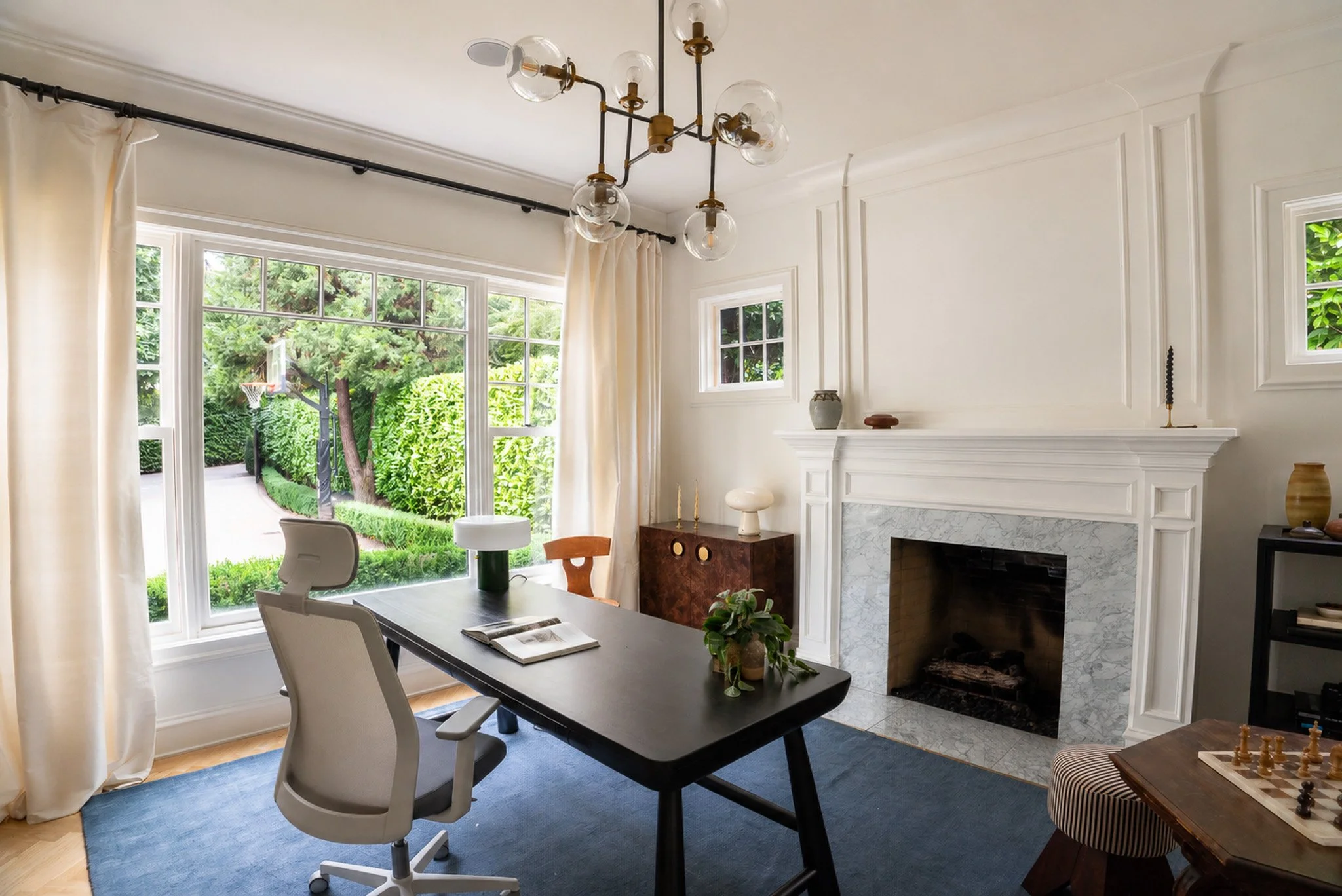 A dining room with large windows showing greenery outside. The room has a black dining table, white chairs, a modern chandelier, and a fireplace with decorative items and small windows.