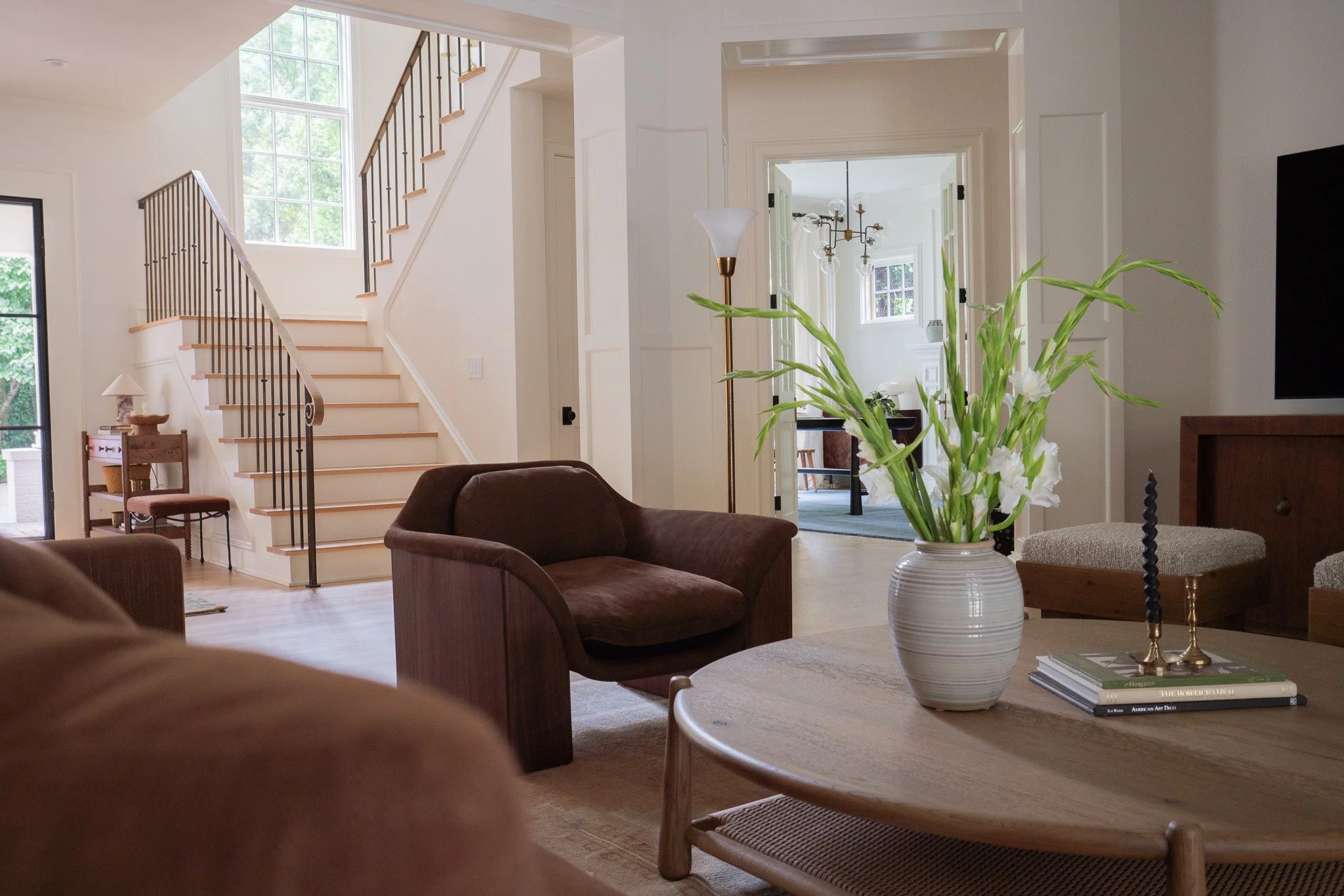 Living room with a dark brown sofa, light wood coffee table with a white vase of white flowers, a stack of books, and various chairs. Large window, staircase, and open doorway visible.