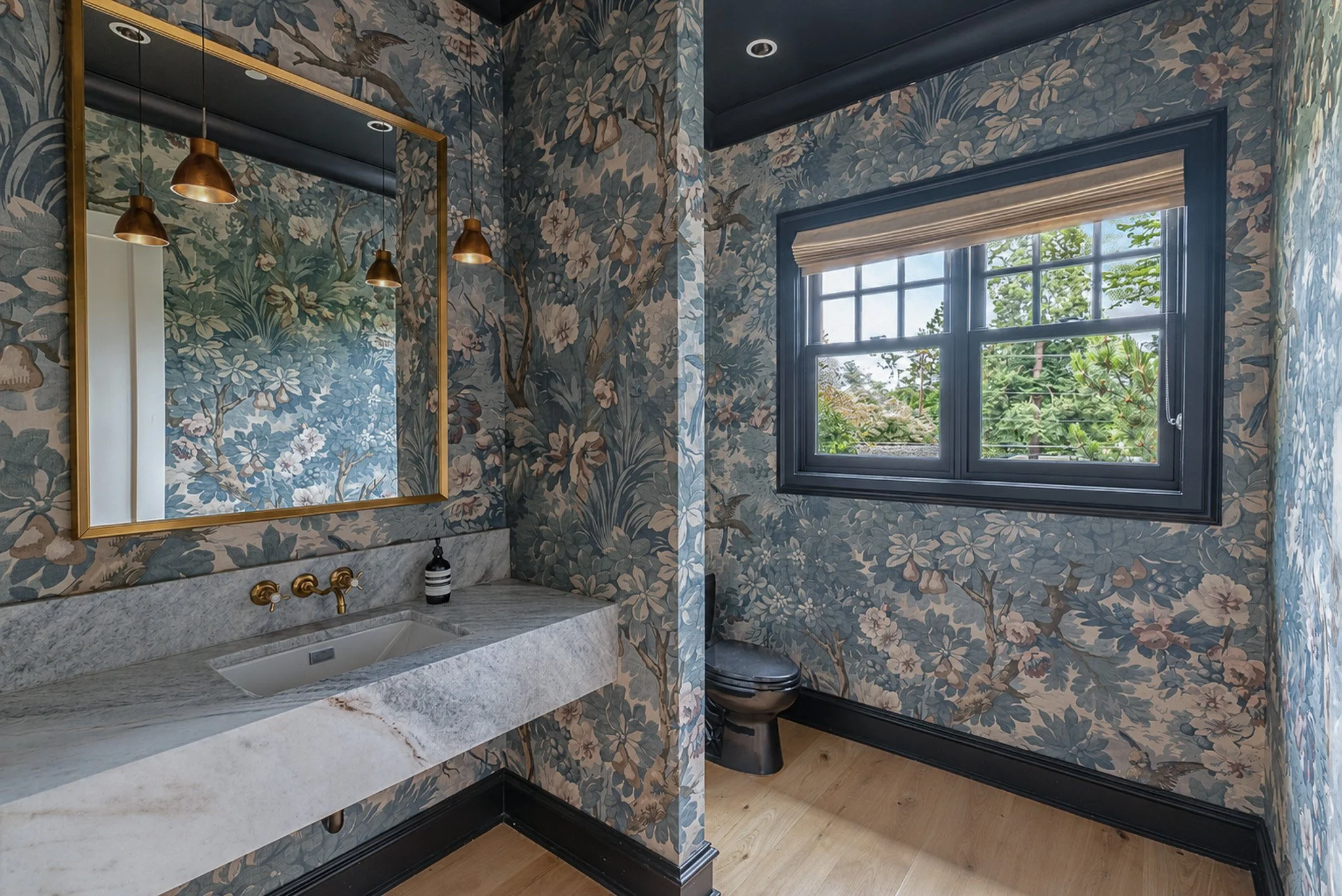 Bathroom with floral-patterned wallpaper, a large mirror with a gold frame, black fixtures, marble countertop with a sink, and a window showing greenery outside.