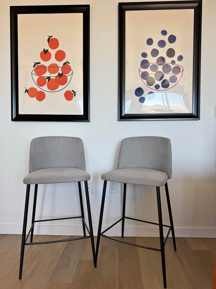Two gray bar stools in front of two framed prints of fruit bowls, one with cherries and the other with blueberries, hanging on a white wall.