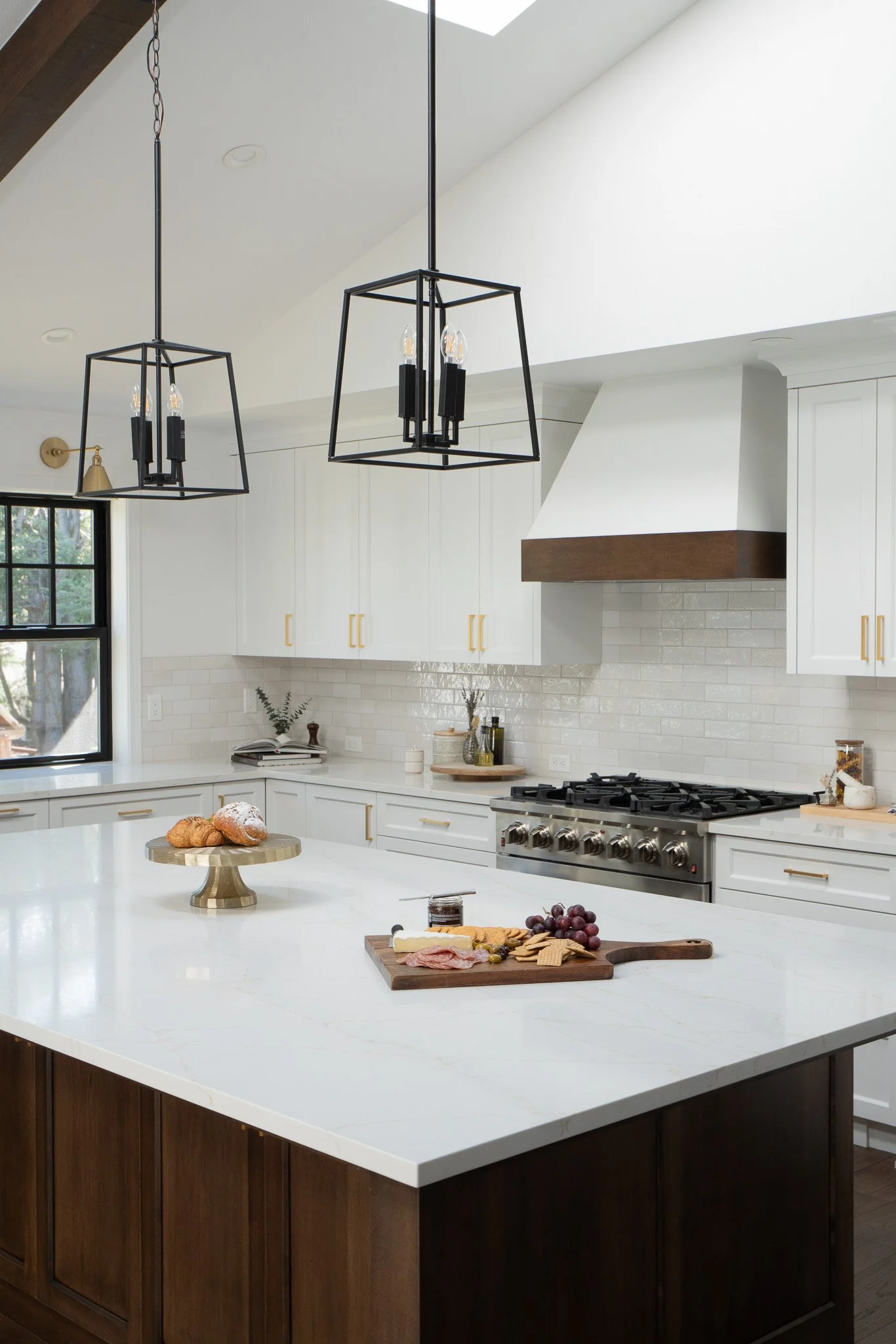 A modern kitchen with white cabinets, a white marble island, black pendant lights, a stove, and various food items on the island and counter.