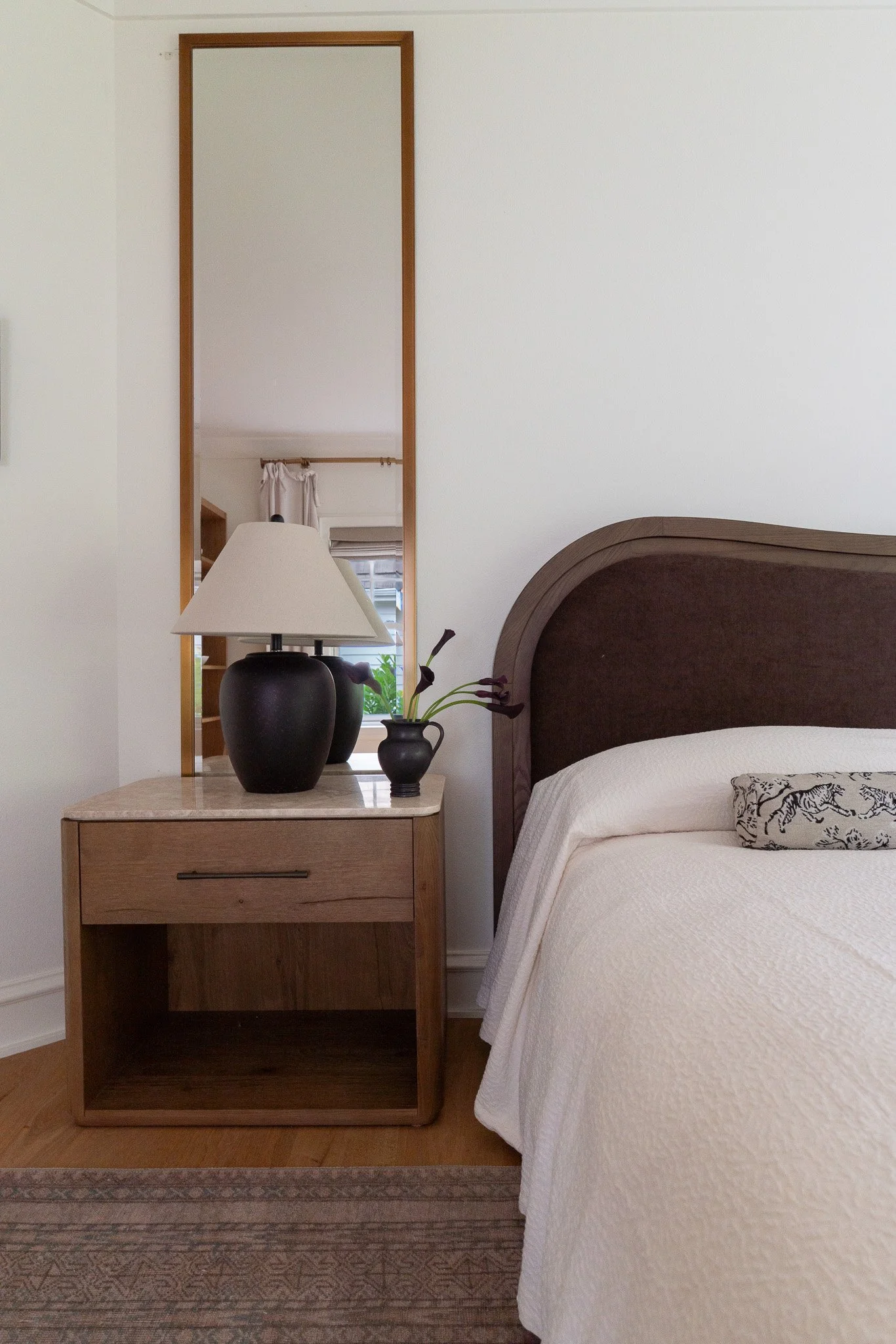 A bedroom with a wooden nightstand, black vases, a lamp with a beige shade, a bed with white bedding, and a mirror reflecting part of the room.