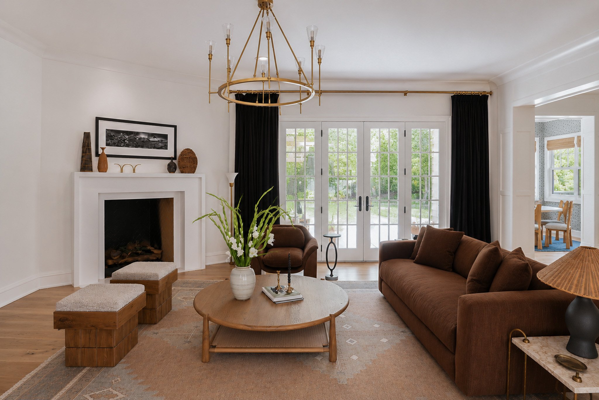 A living room with a large window and door opening to a garden, featuring a brown sofa, two armchairs, a wooden coffee table with a flower vase, and decorative items on the white fireplace mantel, with a chandelier hanging from the ceiling.