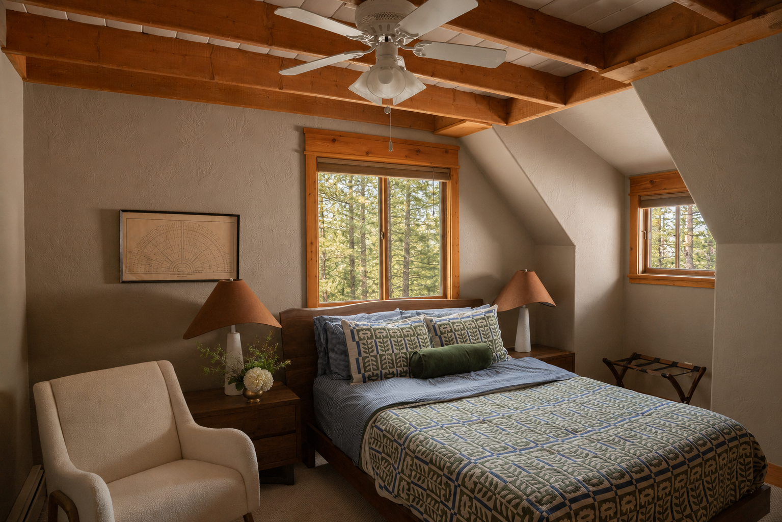 A cozy bedroom with a bed that has patterned bedding and pillows, two wooden nightstands with lamps, a beige armchair, and windows showing trees outside, with a wood-beamed ceiling and a ceiling fan.