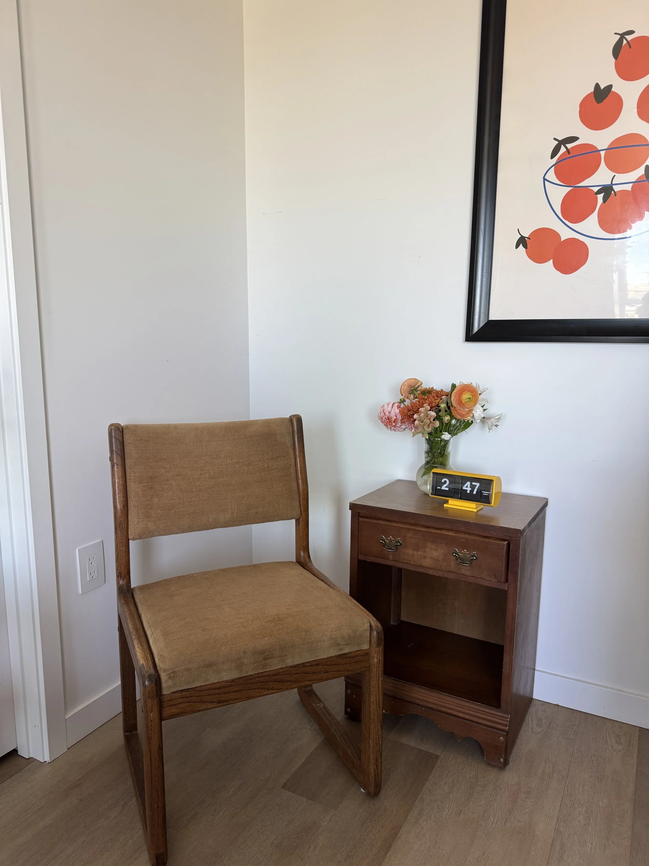 A wooden chair with a cushioned tan seat next to a small wooden nightstand holding a vase of flowers and a digital clock showing 2:47, on a light-colored floor against a white wall with a framed artwork of oranges on the right side.