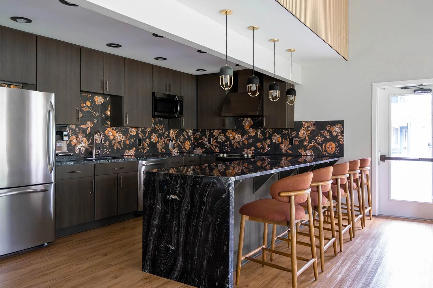 Modern kitchen with dark wood cabinets, floral black backsplash, stainless steel refrigerator, black marble countertop island, and four pink upholstered bar stools in front of a door.