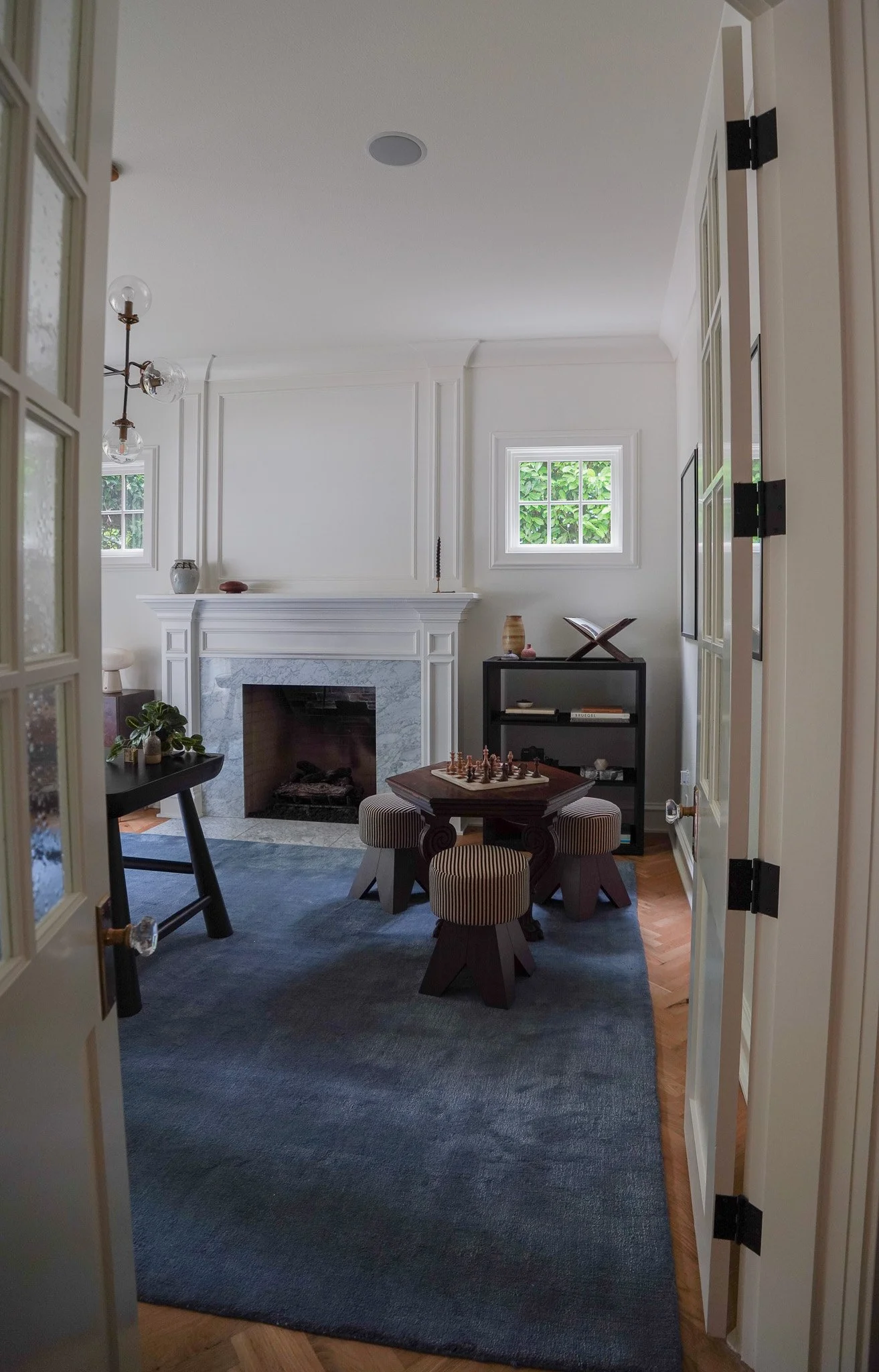 Interior view of a living room with white walls and fireplace, small windows showing greenery, and furniture including a round table with striped stools and a black bookshelf.