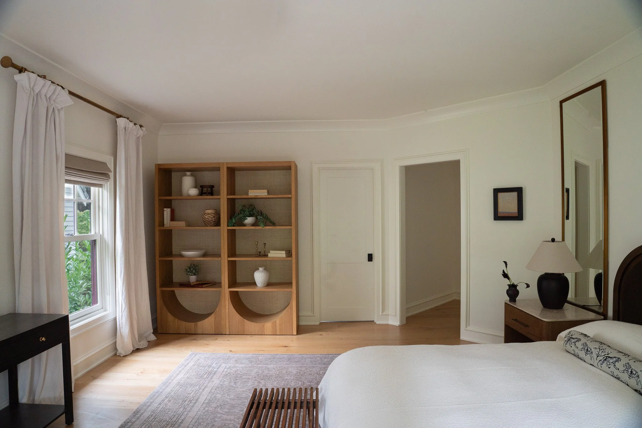 Interior of a bedroom with a window and white curtains, a wooden shelf with decorative items, a part of a bed with a white duvet, a mirror on the wall, and a nightstand with a lamp and a small plant.