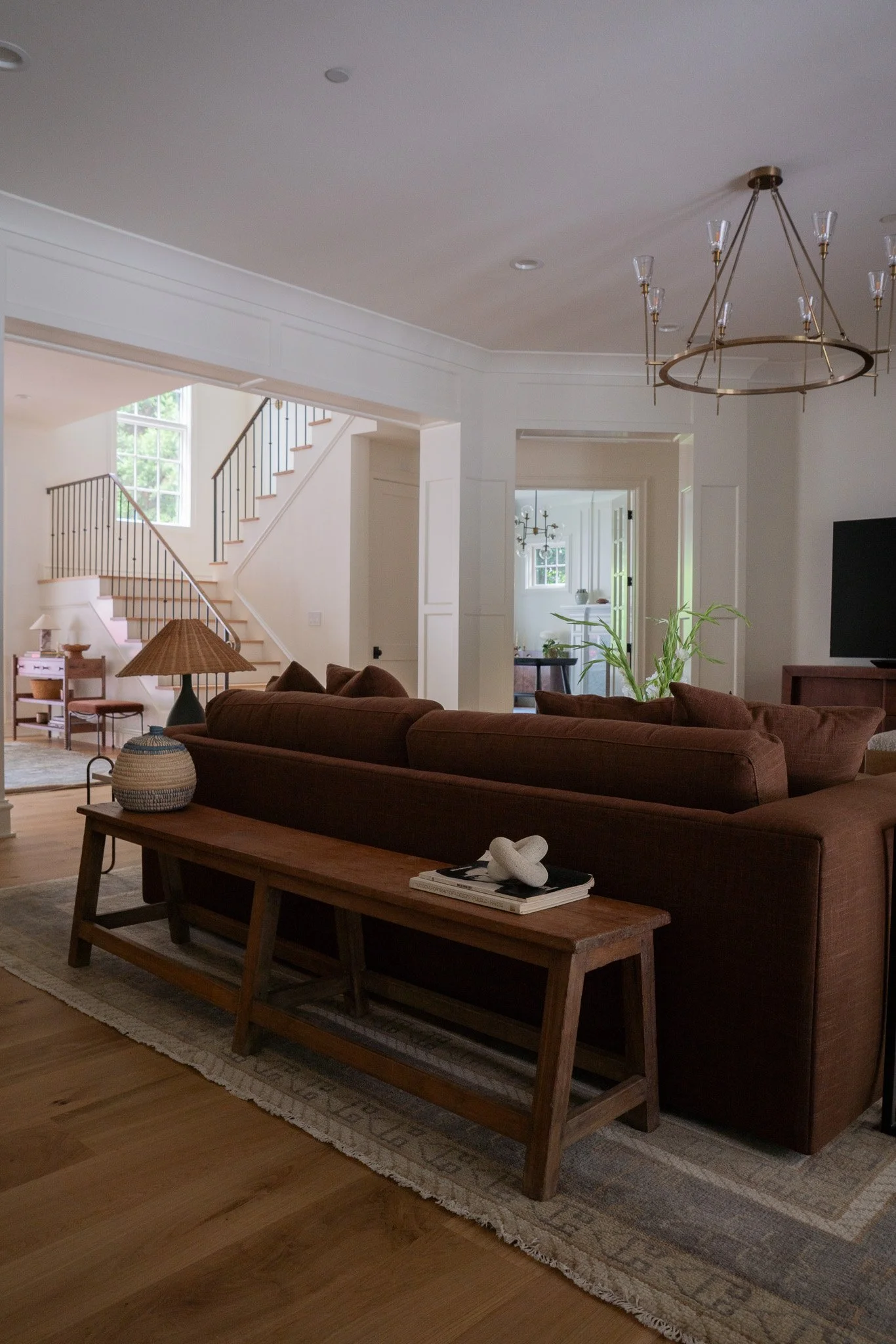 Living room with a brown sofa, wooden coffee table with a basket and books, a decorative sculpture, a rug, and a plaid armchair to the left. In the background are stairs with a black railing, large windows, and a dining area with a chandelier.