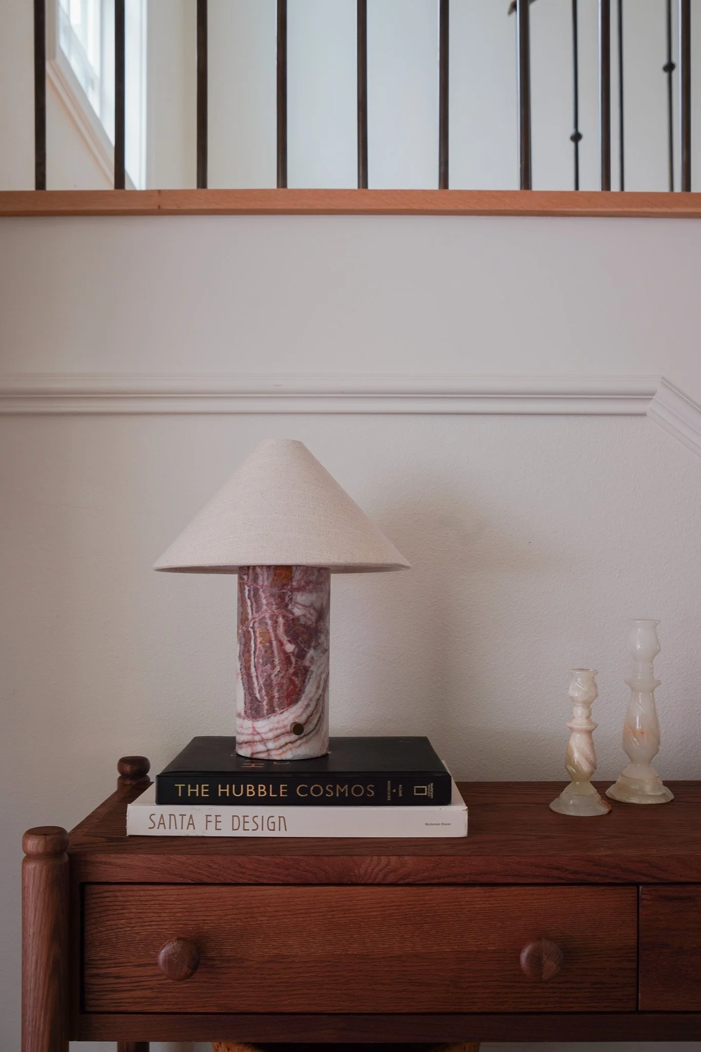 A table lamp with a pink and white marble base and a beige lampshade on a wooden table. Beneath the lamp are two stacked books titled 'The Hubble Cosmos' and 'Santa Fe Design'. To the right of the lamp, there are two small, cream-colored decorative v