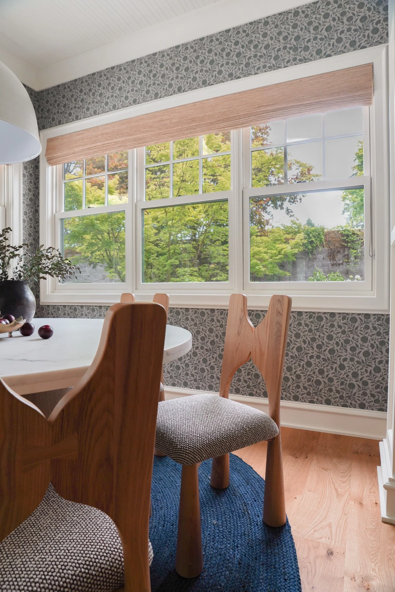 A dining room interior with a large window showing trees outside, wooden chairs, a table, and patterned wallpaper.