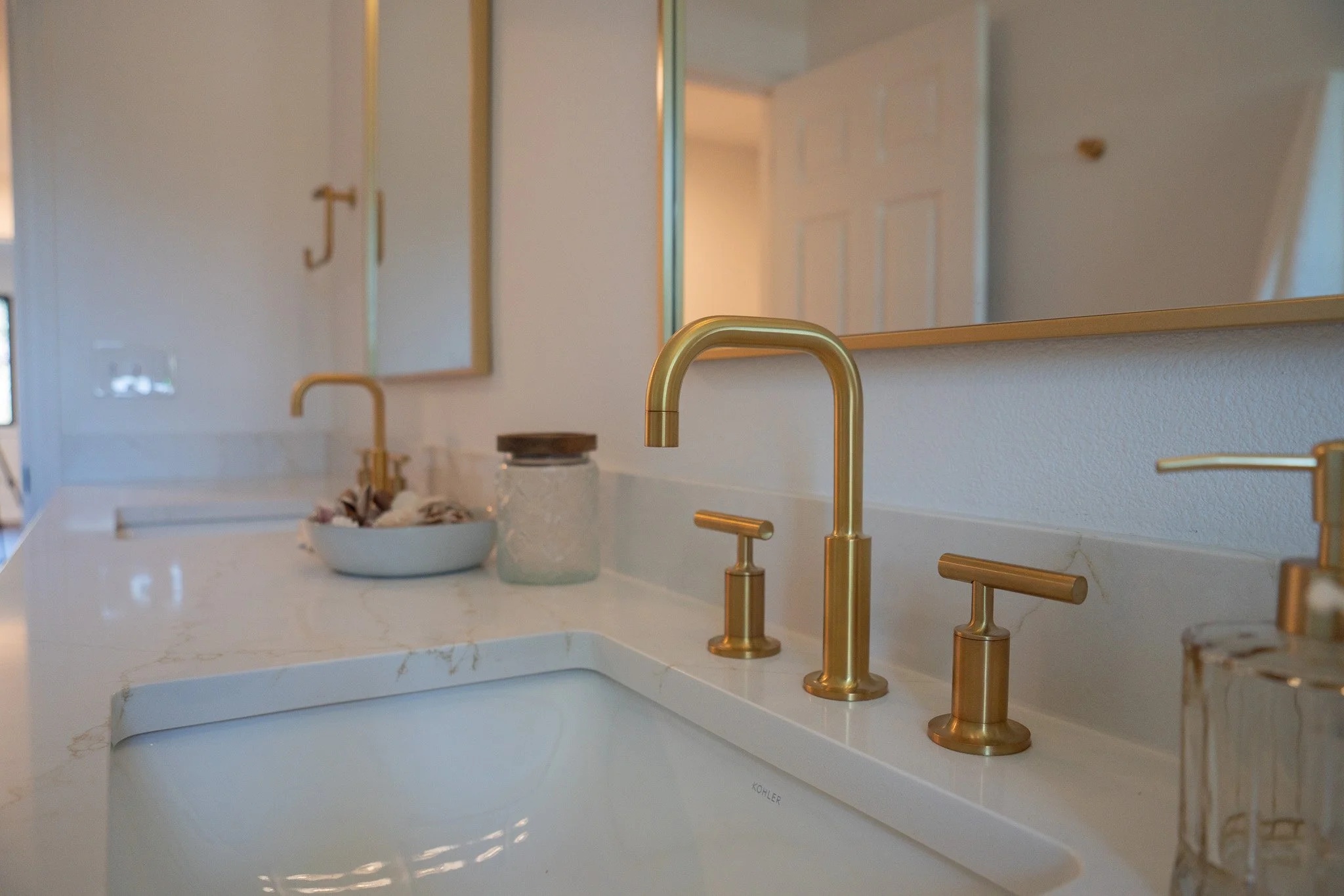 A bathroom sink with two gold faucets and a mirror above it. There is a white countertop with a bowl and jars. The background shows a door and light-colored walls.