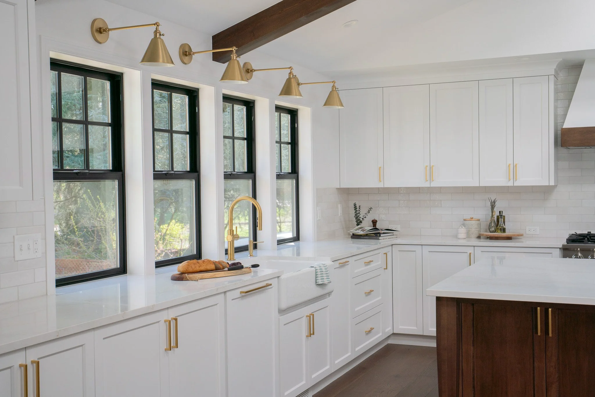 Modern kitchen with white cabinets, black window frames, a white quartz countertop, gold fixtures, and a dark wood kitchen island.