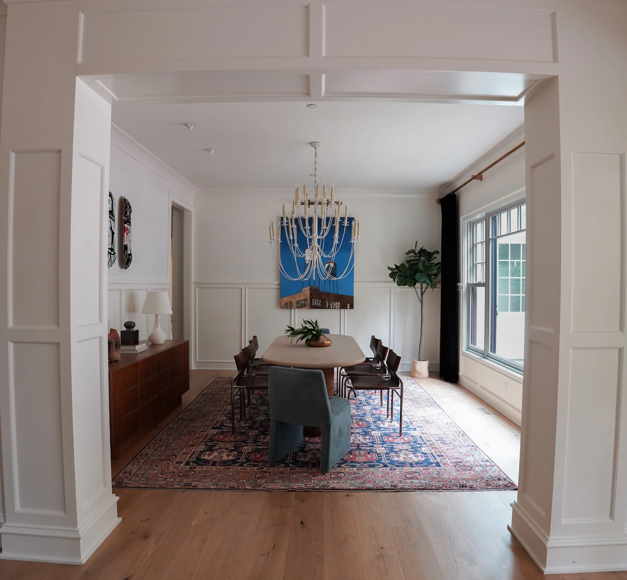 Interior view of a dining room with a rectangular table surrounded by six chairs. There is a large rug underneath the table, and a chandelier hanging above. The room has white wainscoting, a large window with black curtains on the right, and a painti