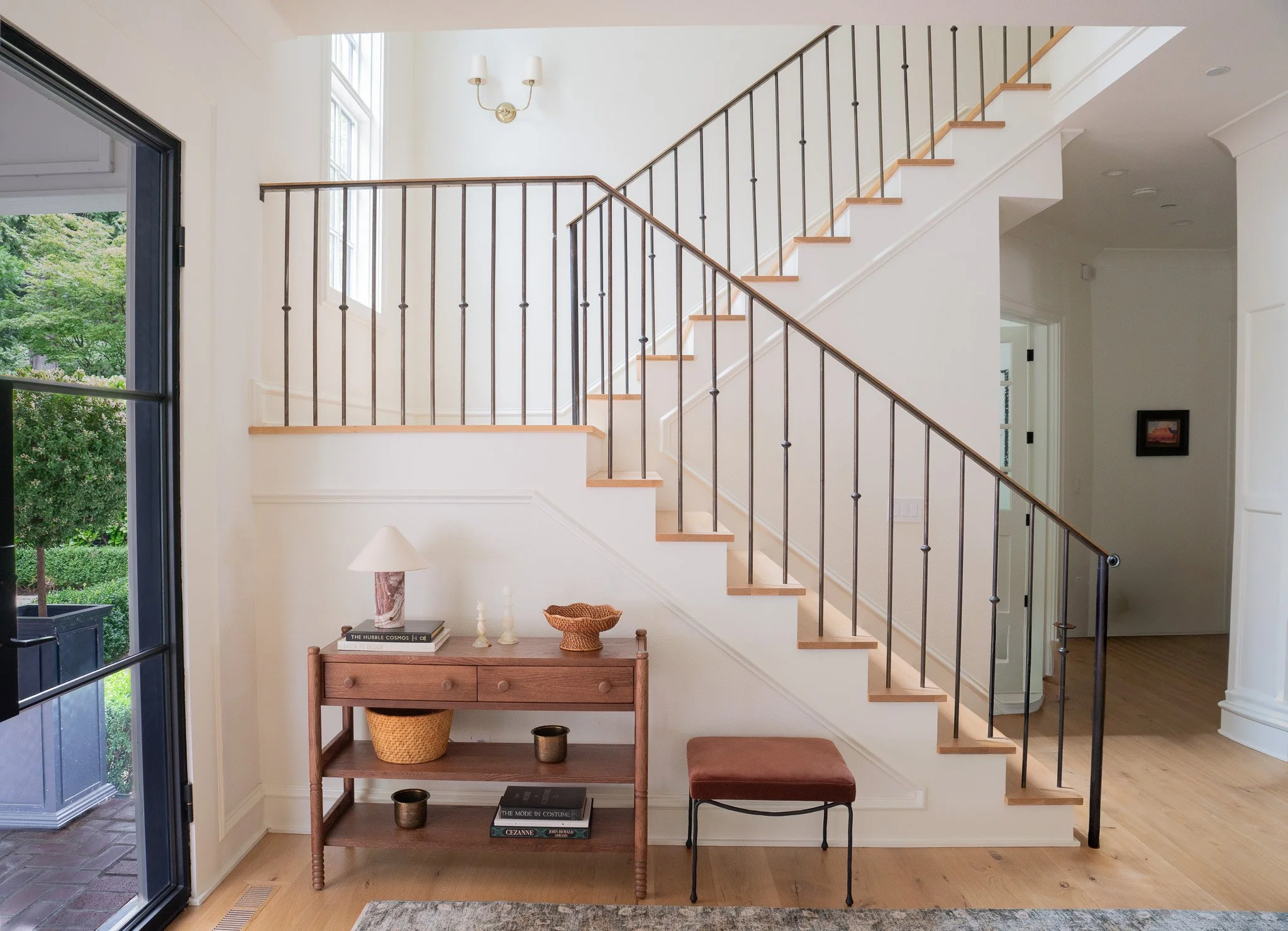 Interior view of a house showing stairs with wooden steps and black metal railing, white walls, a small wooden table with books and decorative items, a window on the upper left, and a sliding glass door open to greenery outside.