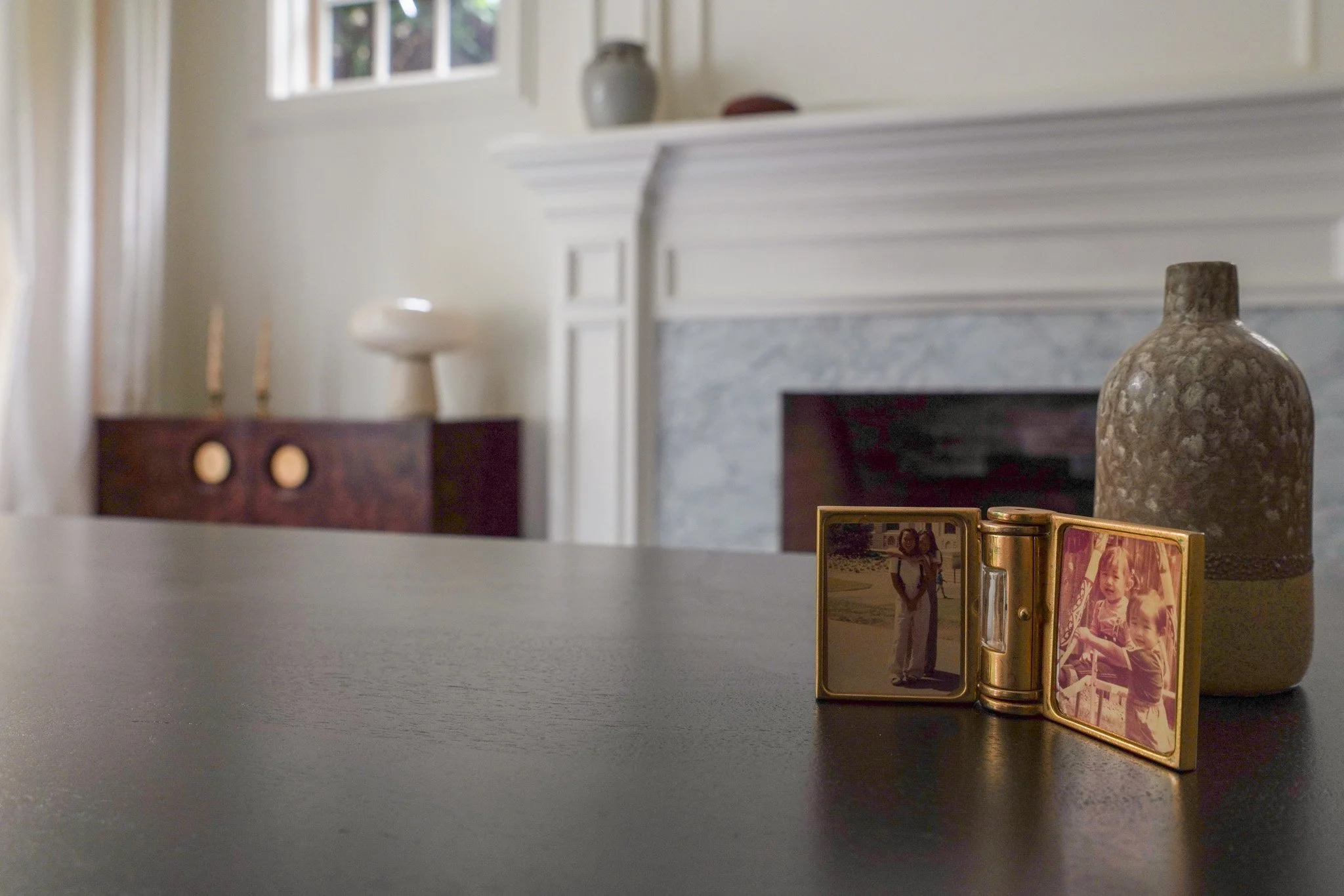 Close-up of small, vintage-style photo frame with pictures of a woman and children, along with a gold-colored container, on a black surface. In the background, a marble fireplace and decorative vases are visible in a room with a window.