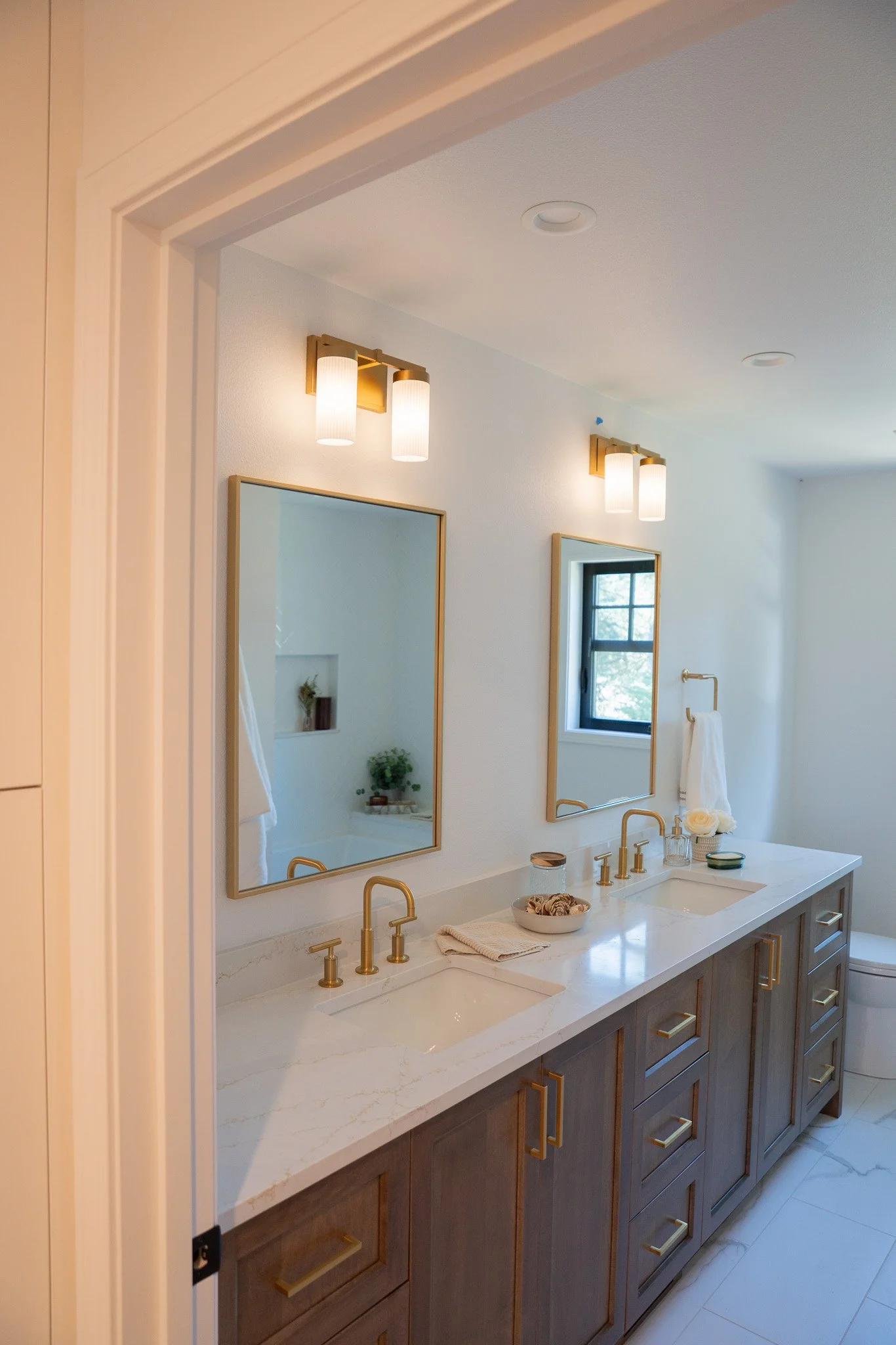 A modern bathroom vanity with a white marble countertop, two undermount sinks with gold faucets, and two mirrors framed in gold. Above the vanity, there are two wall-mounted light fixtures with cylindrical shades. The vanity has wooden cabinets with 