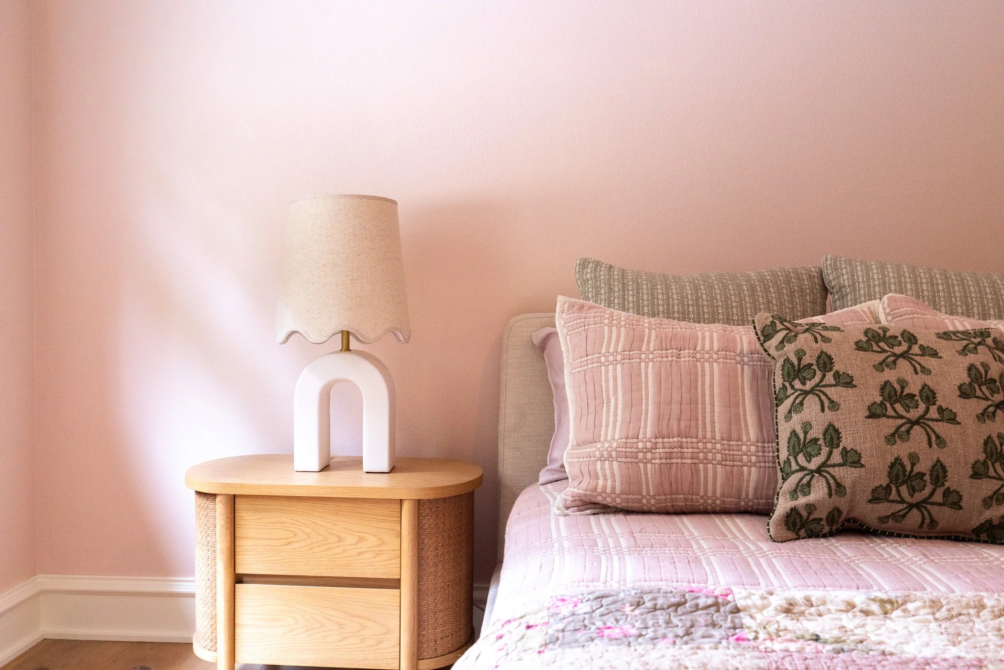A bedroom corner with a pink wall, a wooden bedside table, a white modern lamp with a beige lampshade, and a bed with pink and green floral and striped pillows.
