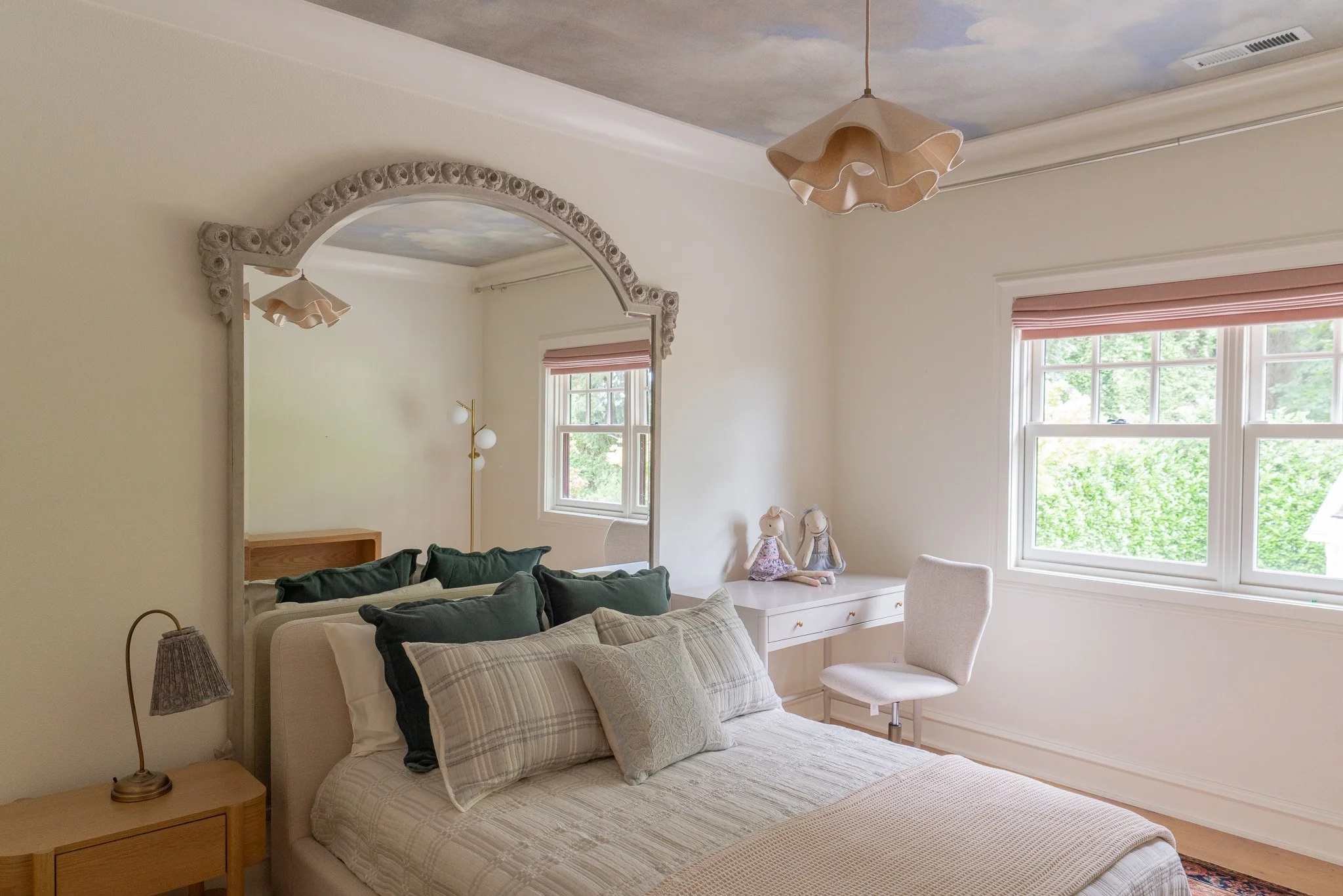 A bedroom with a white bed, multiple pillows, and a bedside table with a lamp. There are two dolls on a white dresser near the window. The room has a ceiling with a sky mural and a decorative archway with floral details.