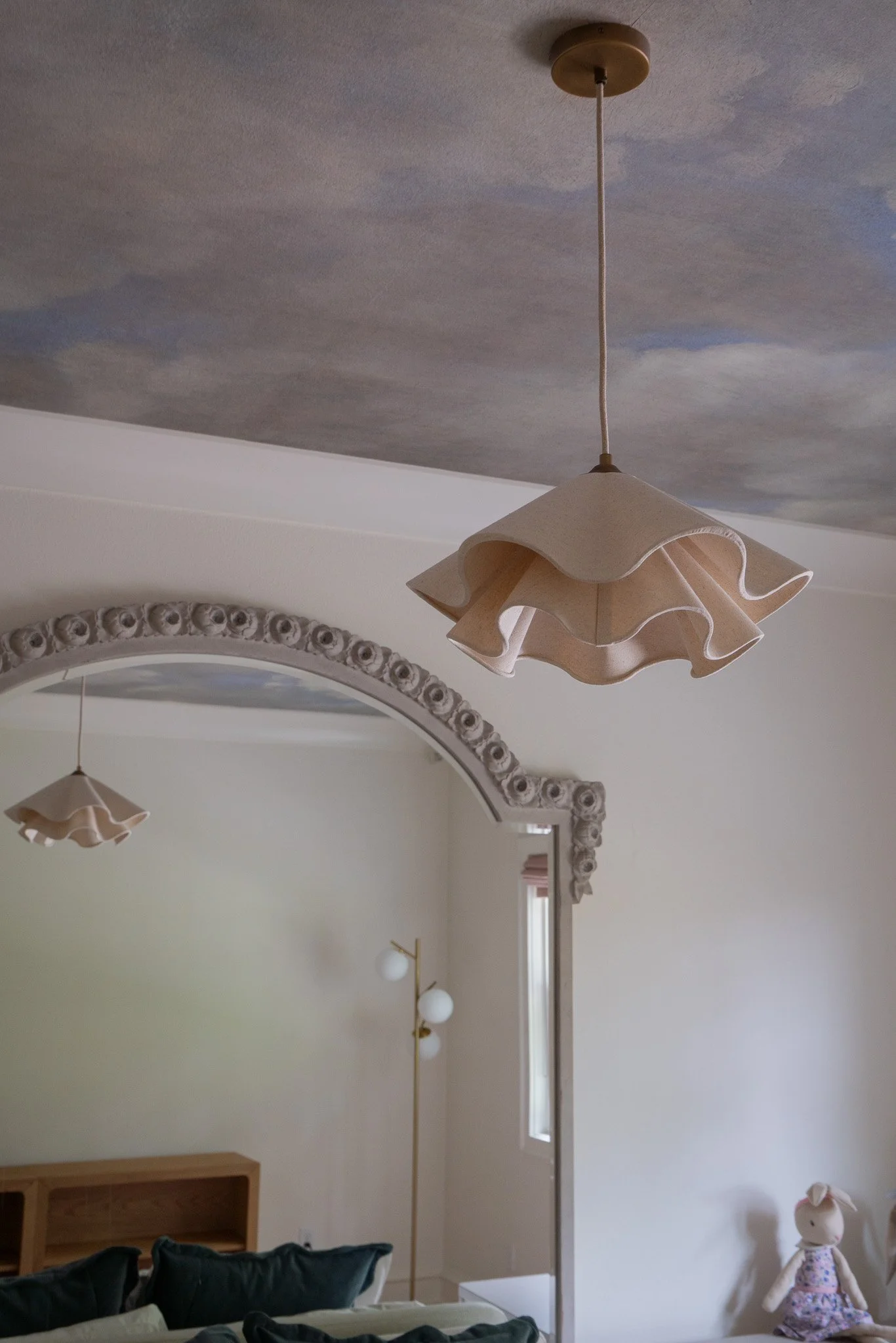 Living room ceiling with a large, beige flower-shaped pendant light and a mural of a cloudy sky on the ceiling. Part of a mirror with decorative frame, a window, a pair of white globe wall sconces, a doll sitting on a chair, and a bookshelf are visib