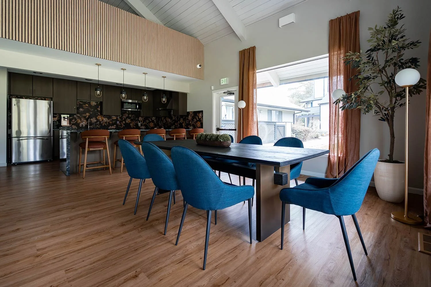 Modern dining area with a large black table surrounded by blue chairs, natural wood flooring, large window with orange curtains, potted tree, and a kitchen in the background with dark cabinetry, a stainless steel refrigerator, and a breakfast bar with brown stools.