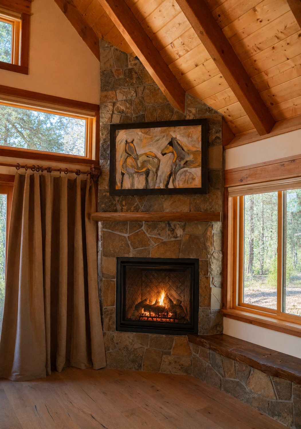A cozy living room corner with a stone fireplace, a painting of two horses above it, and large windows showing a wooded outdoor scene, with wooden walls and ceiling.