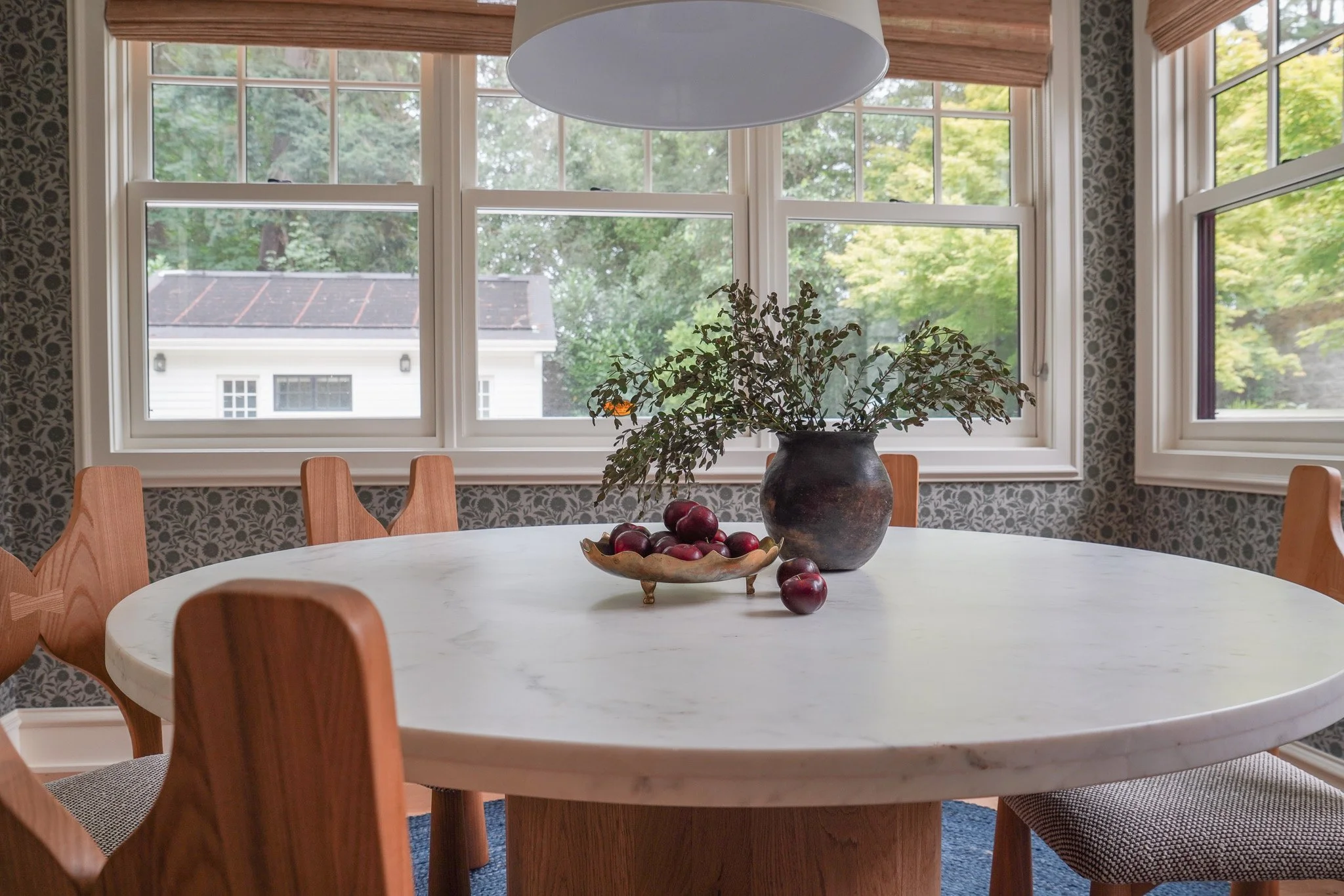 A round white dining table with a ceramic bowl of red apples and a large vase with greenery inside. Wooden chairs surround the table. Large windows with blinds let in natural light, showing trees outside. A white ceiling light hangs above the table. 